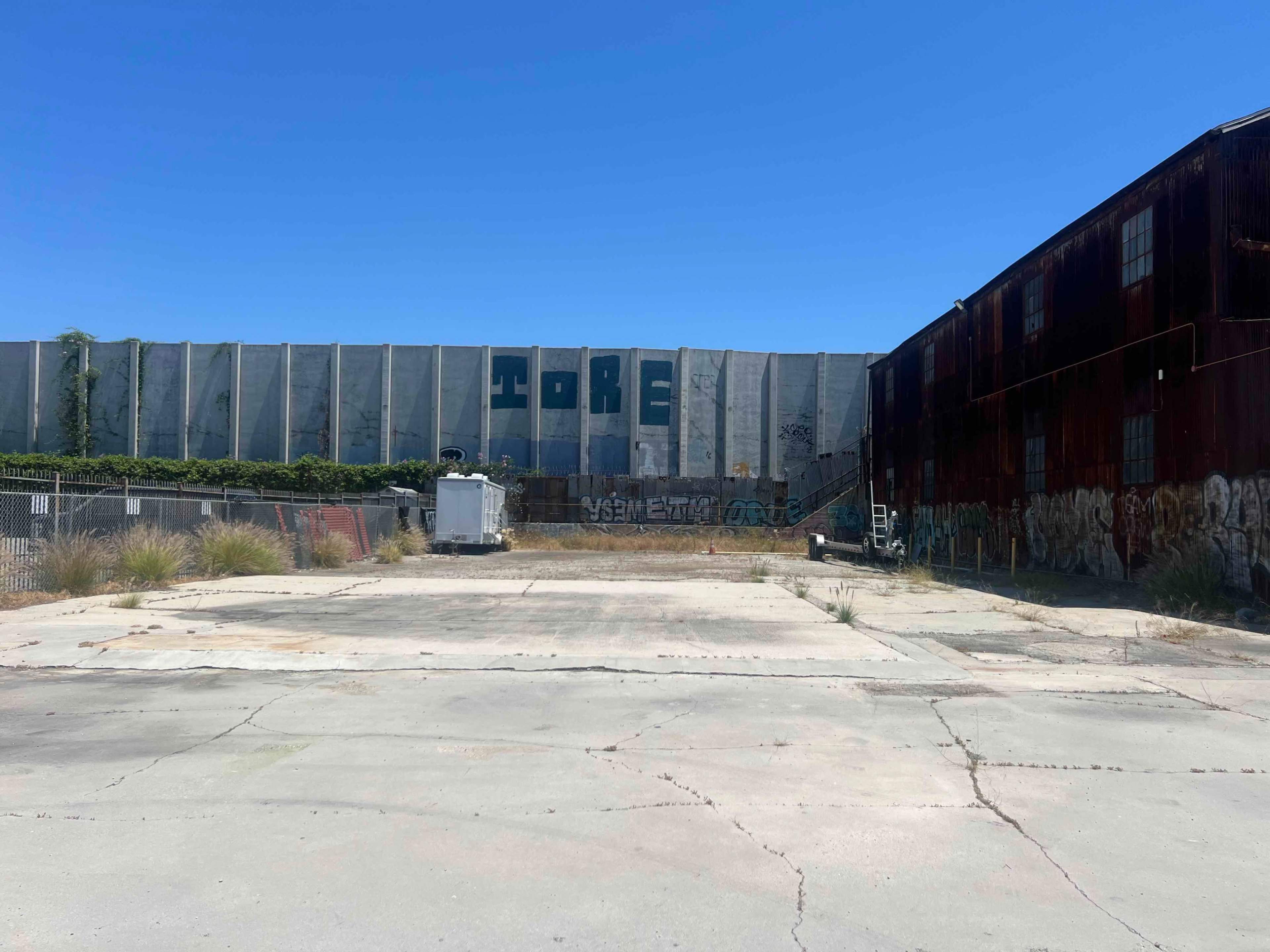 An empty lot in front of a large, graffiti-covered industrial building with a blue sky above.