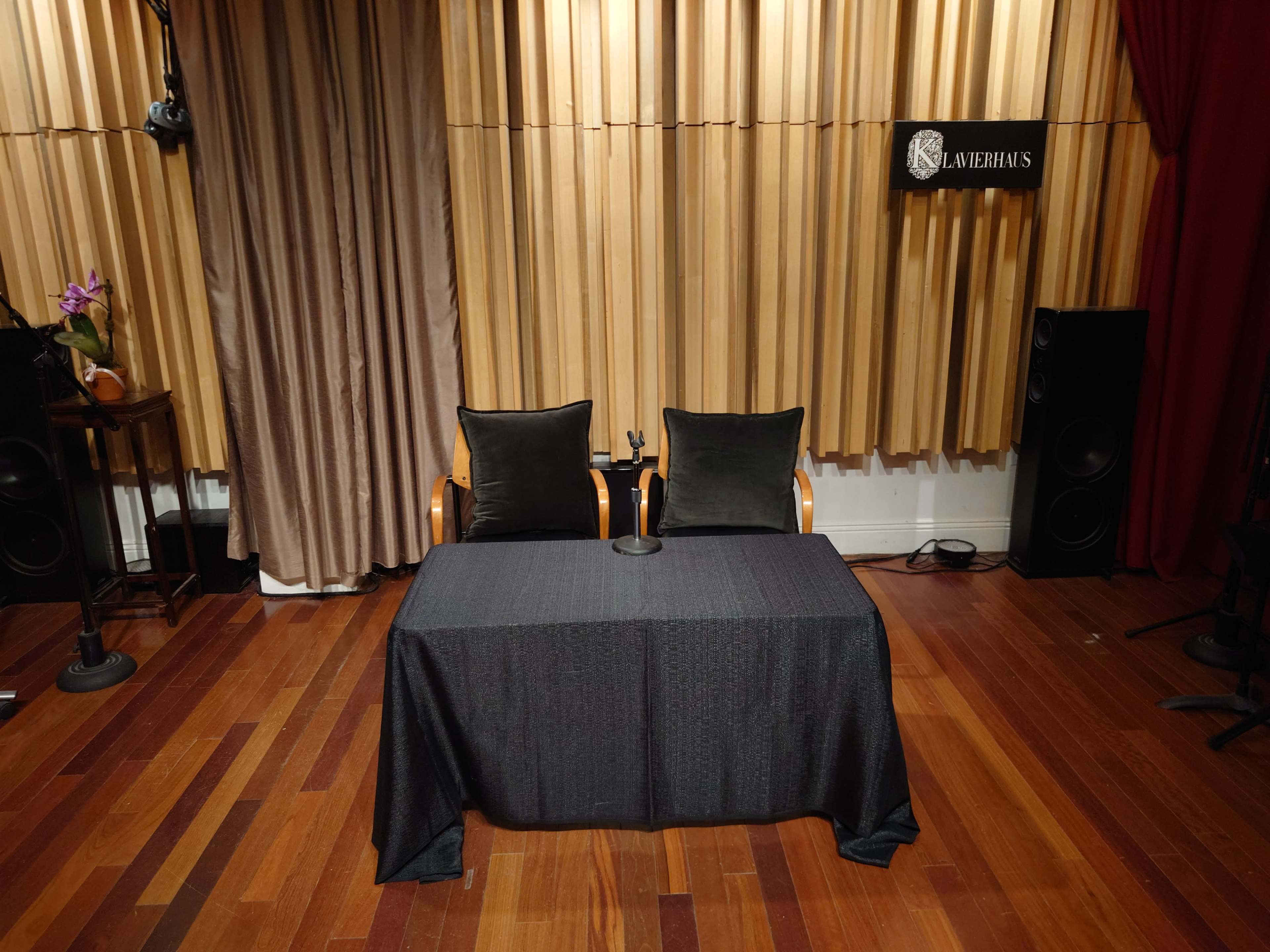 The image shows a stage setup with two chairs and a table covered in a dark cloth, surrounded by sound equipment and draped curtains.