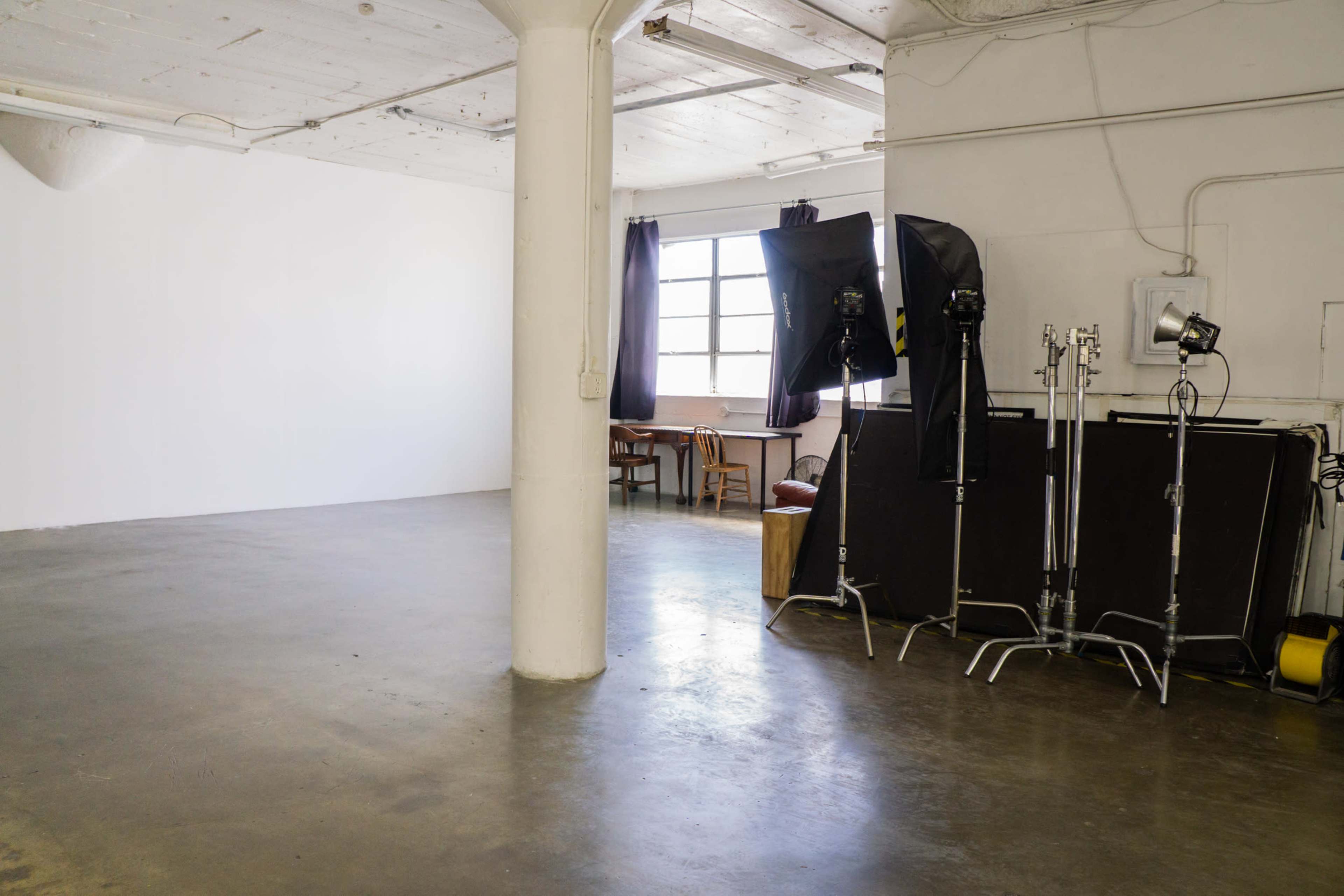 The image shows a spacious indoor area with a concrete floor, a large window, and photography lighting equipment set up against one wall.