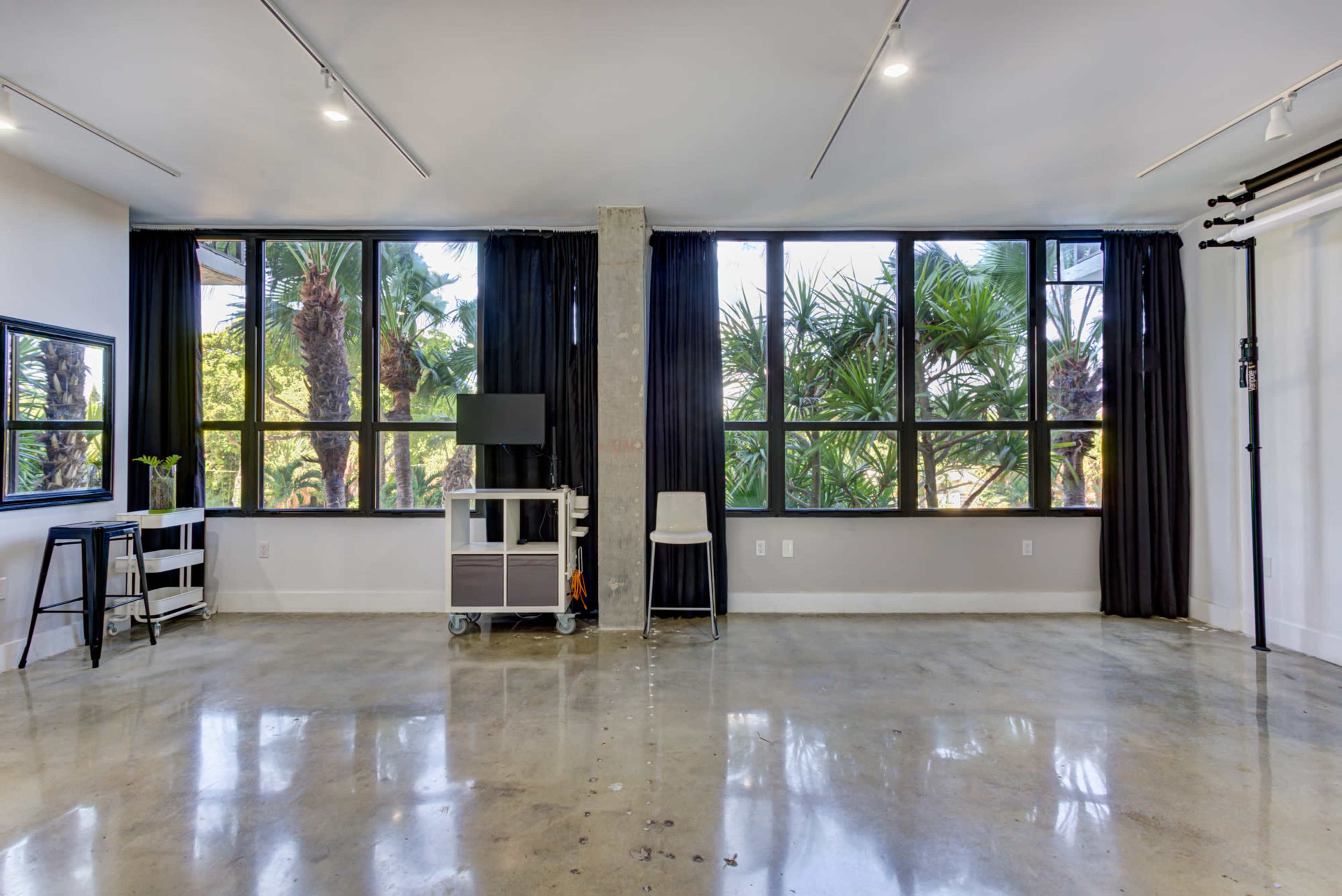 A spacious room with large windows revealing palm trees outside, featuring polished concrete floors and minimal furnishings.