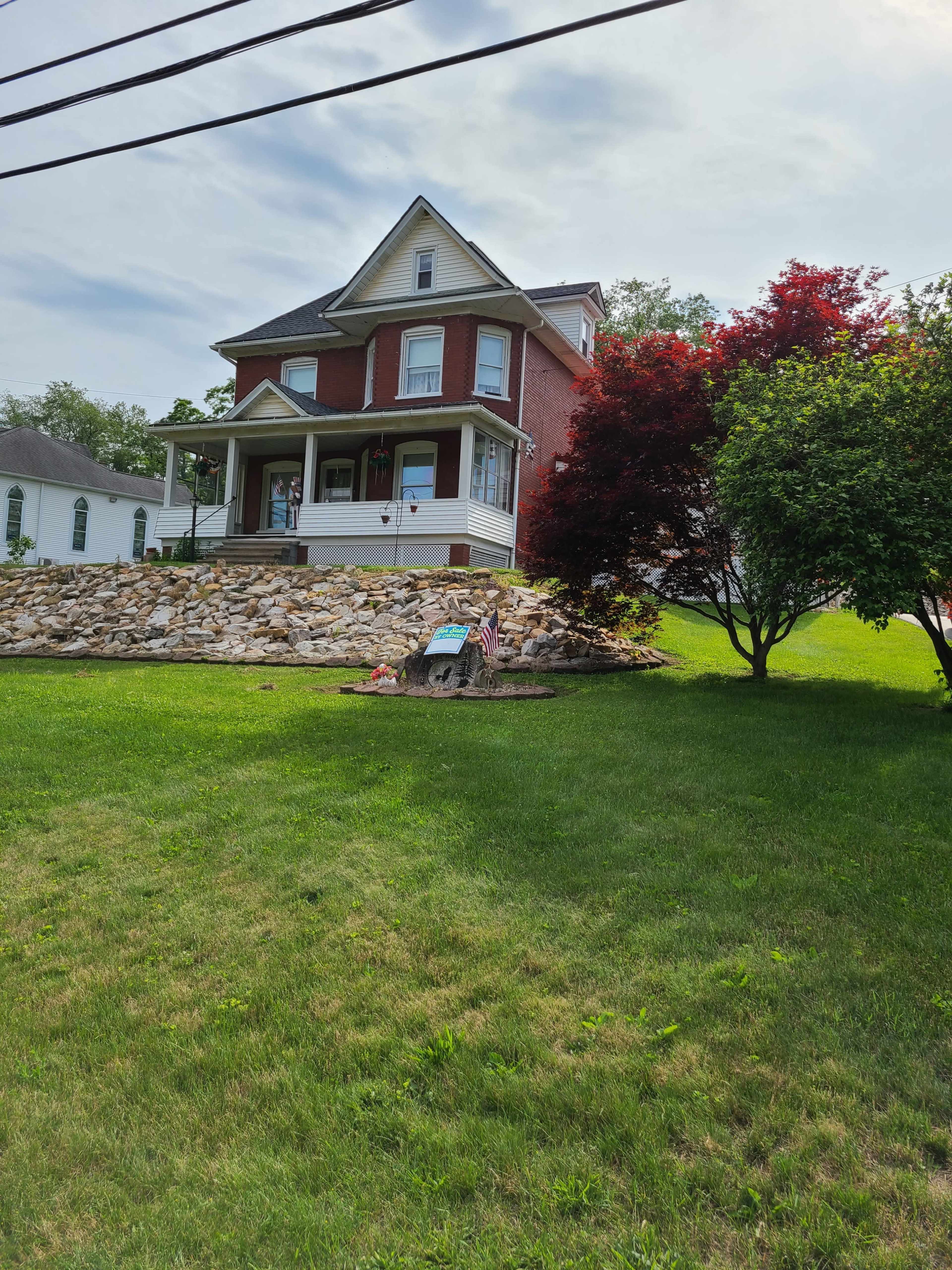 A two-story brick house with a wraparound porch is situated on a grassy lawn, accompanied by a rock landscaping feature and a small garden.