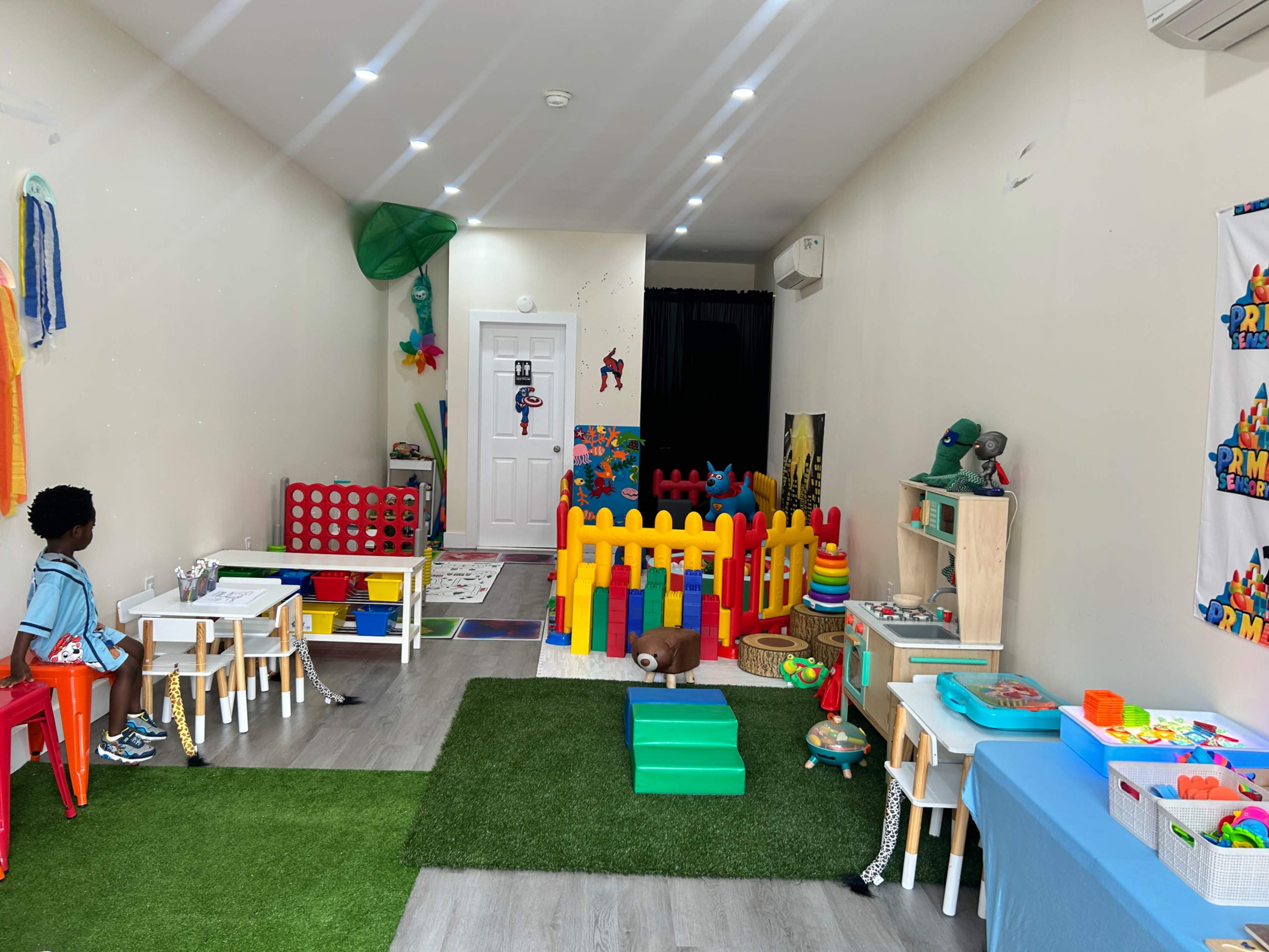 A colorful playroom features a fenced area with toys, small tables and chairs, and artificial grass flooring.