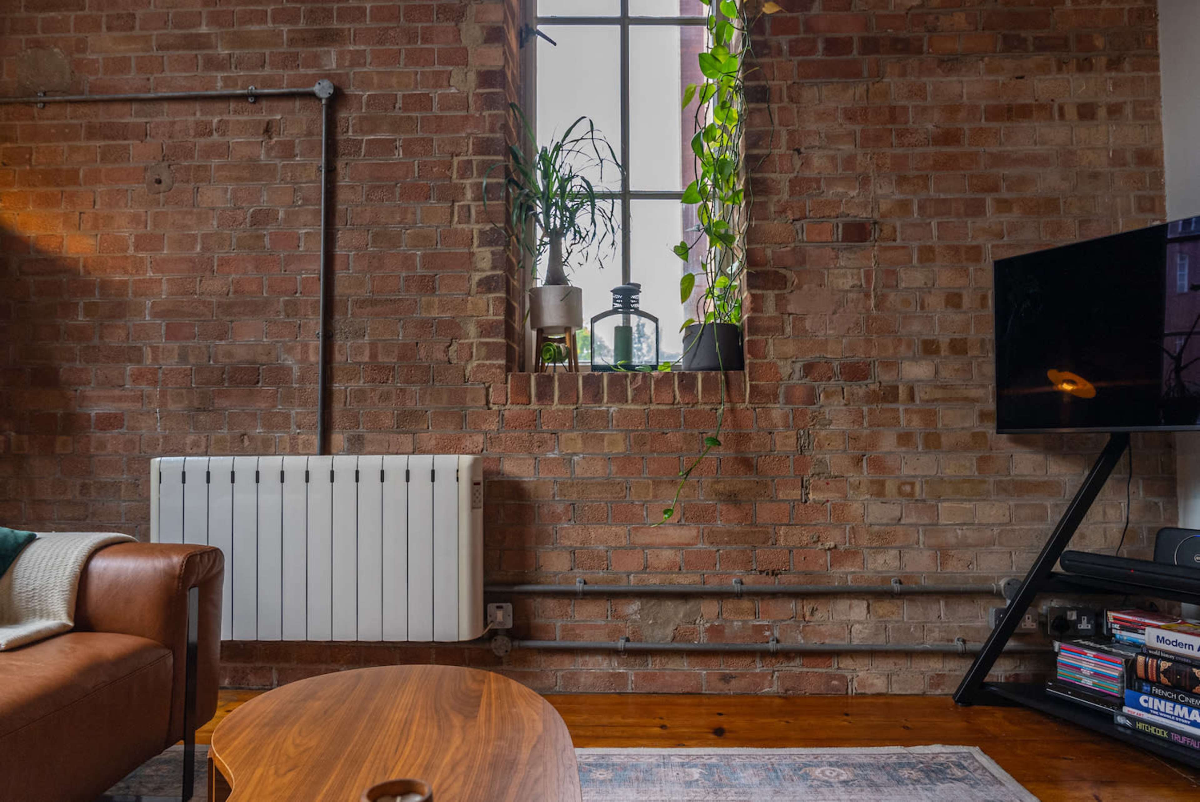 A cozy living space with a leather sofa, a wooden coffee table, a television, and a brick wall featuring a window with potted plants.