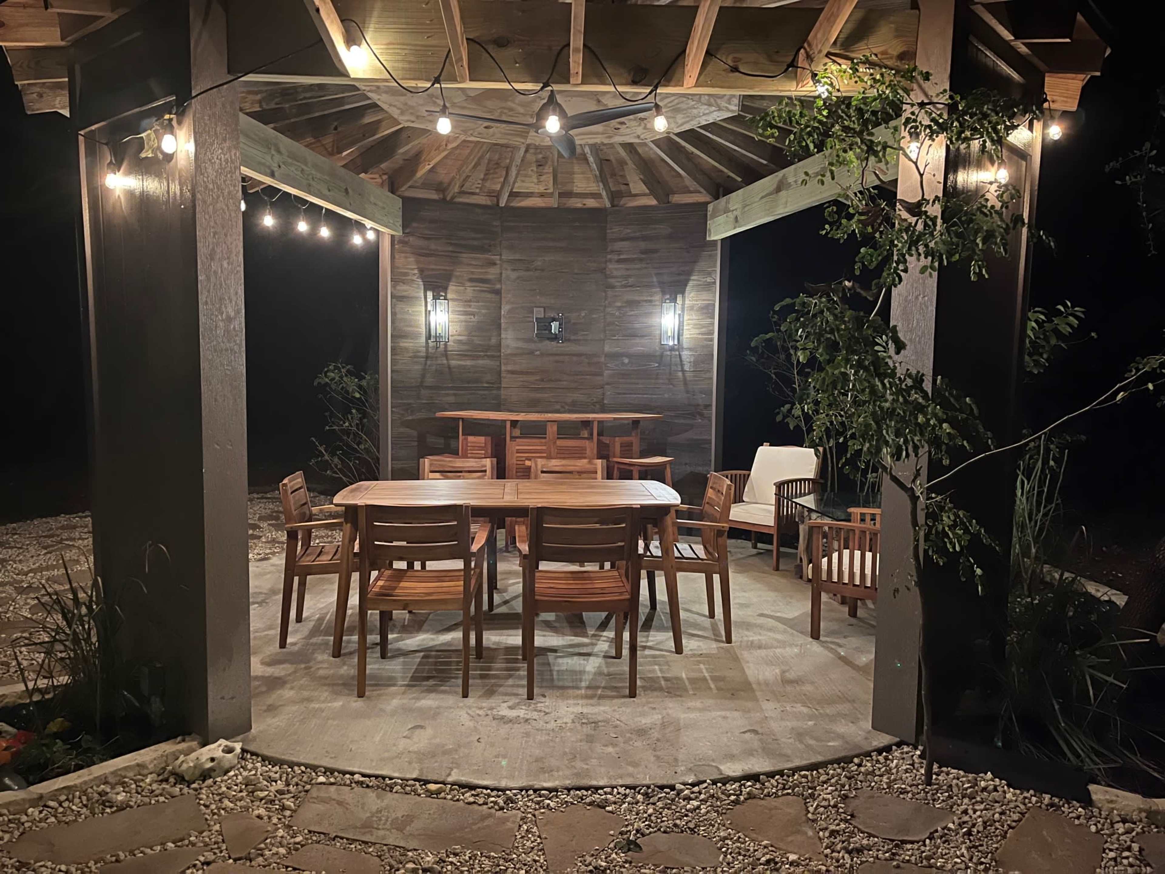 A wooden gazebo with string lights contains a large dining table surrounded by chairs and small seating areas.