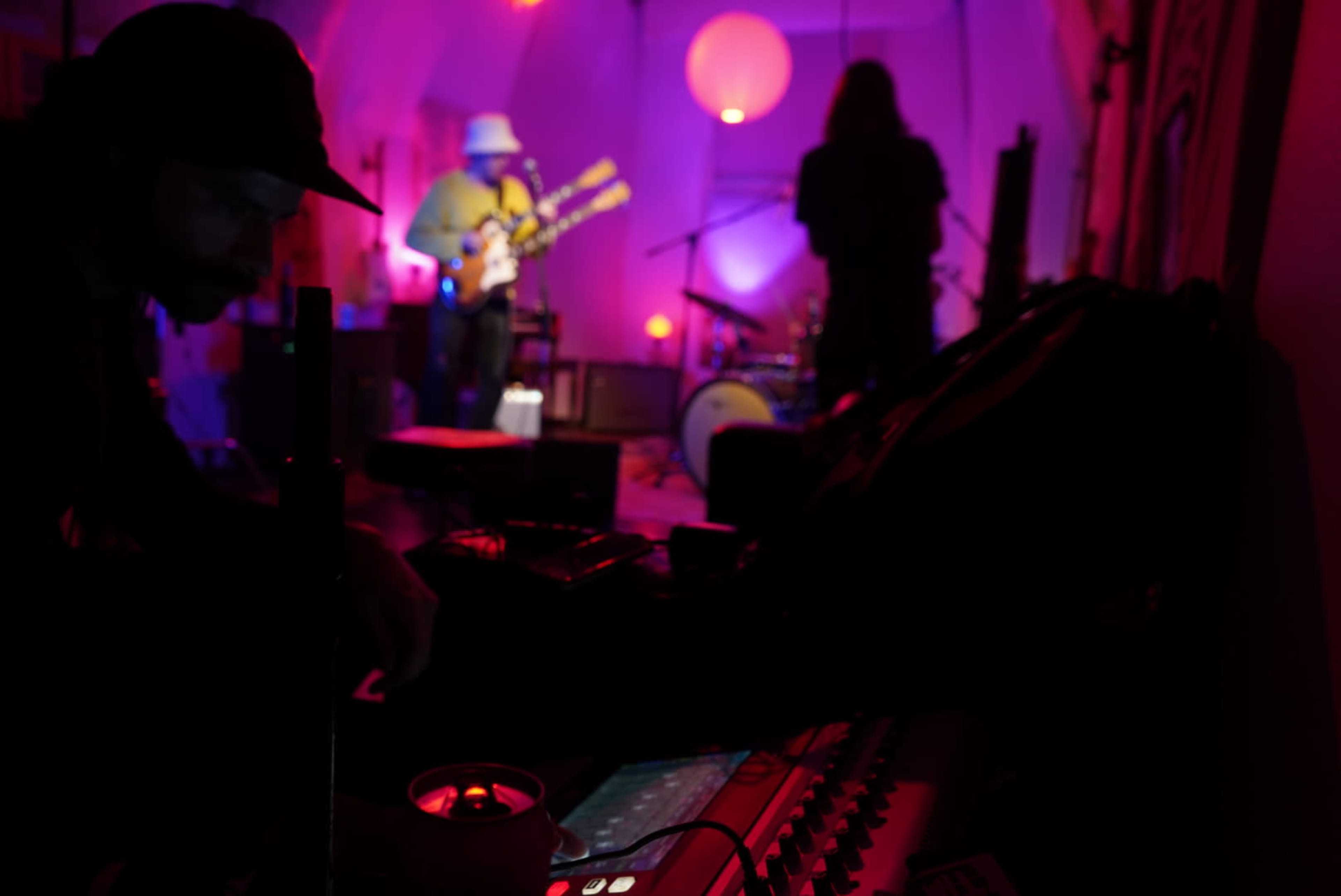 A musician plays guitar on stage while another person adjusts equipment in a dimly lit room.