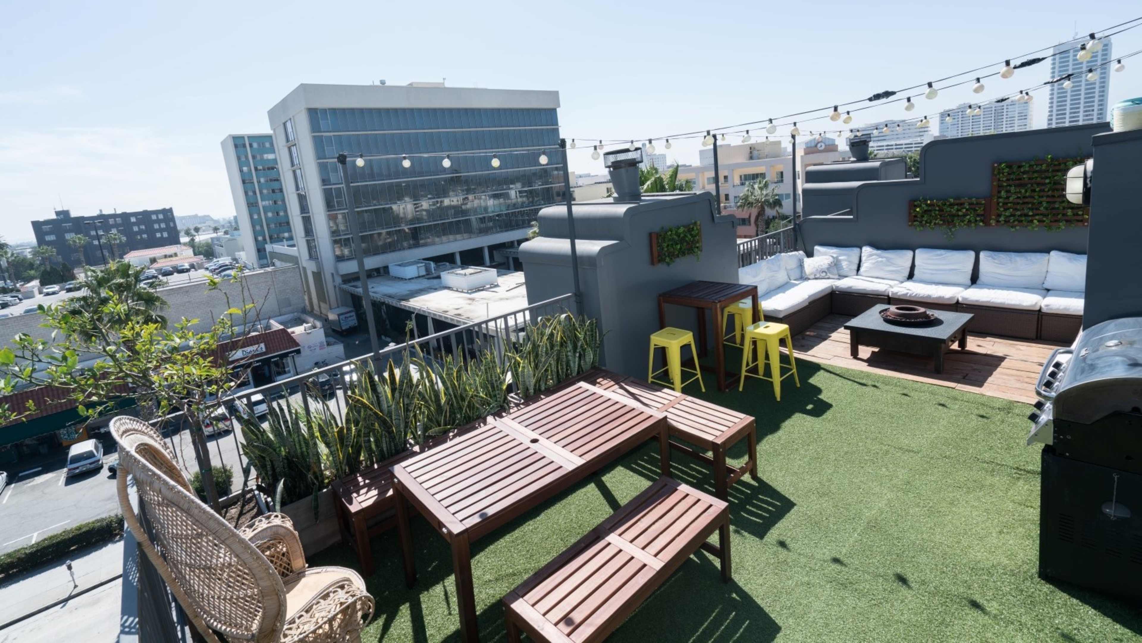 A rooftop terrace with seating areas, wooden tables, and greenery under a clear blue sky.