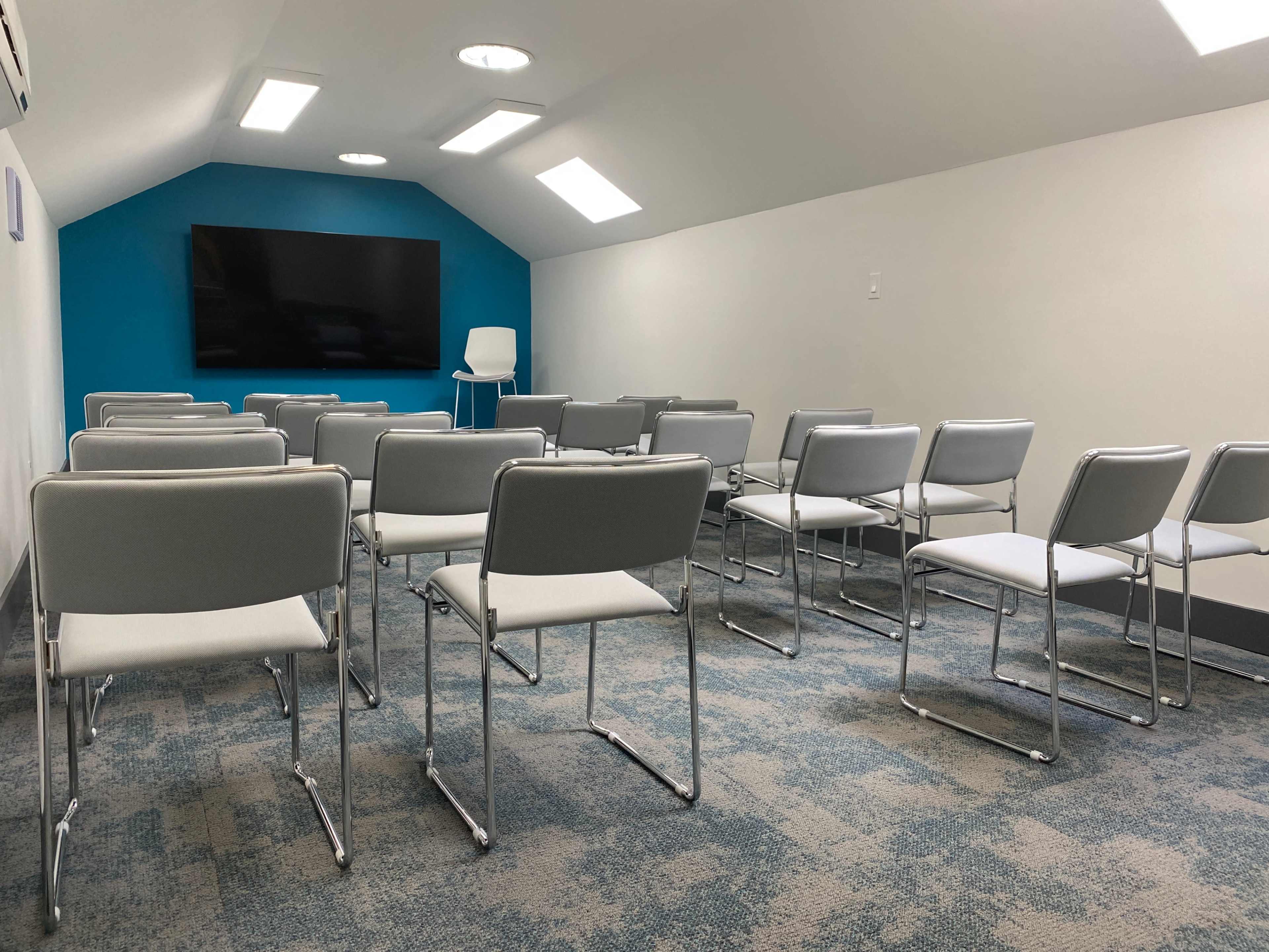 A small meeting room with rows of gray chairs facing a wall-mounted television and a white chair in the corner.