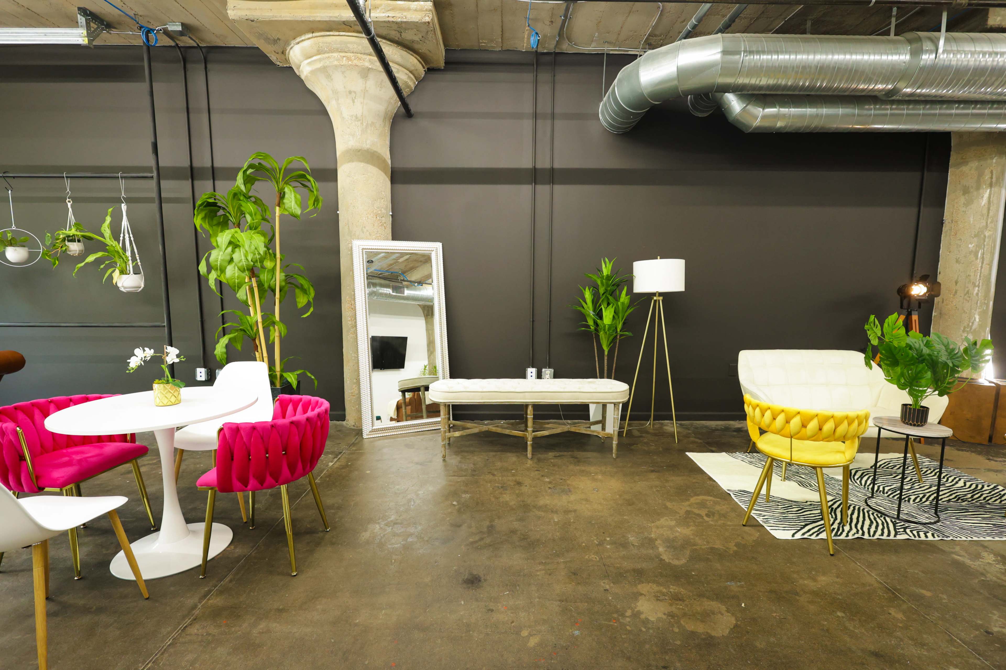 The image shows a modern, minimalist interior space featuring a round dining table with pink chairs, a white bench, a yellow accent chair, a floor lamp, and various plants against a dark wall.