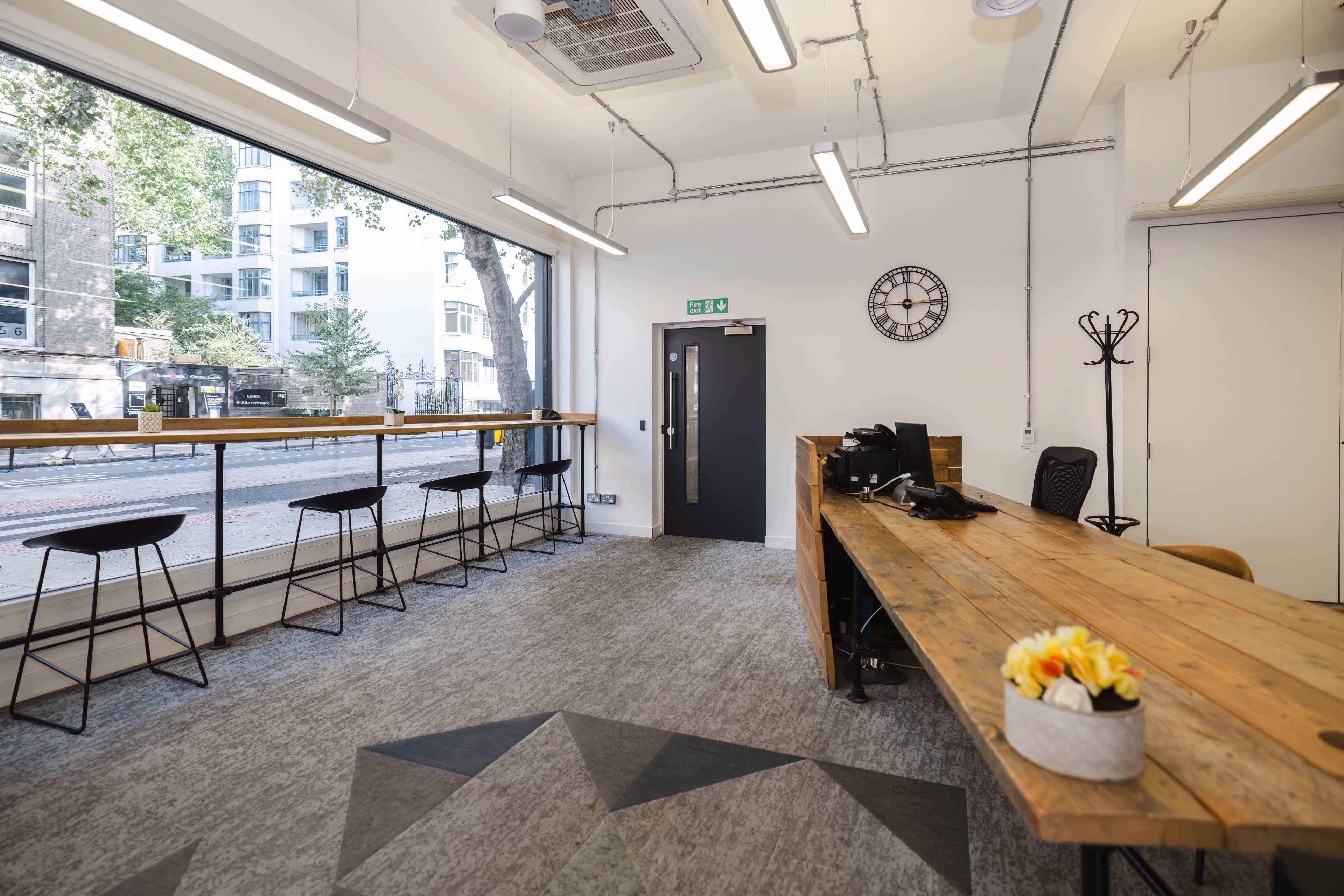 A modern office space with a wooden reception desk, a seating area with bar stools overlooking the street, and large windows letting in natural light.