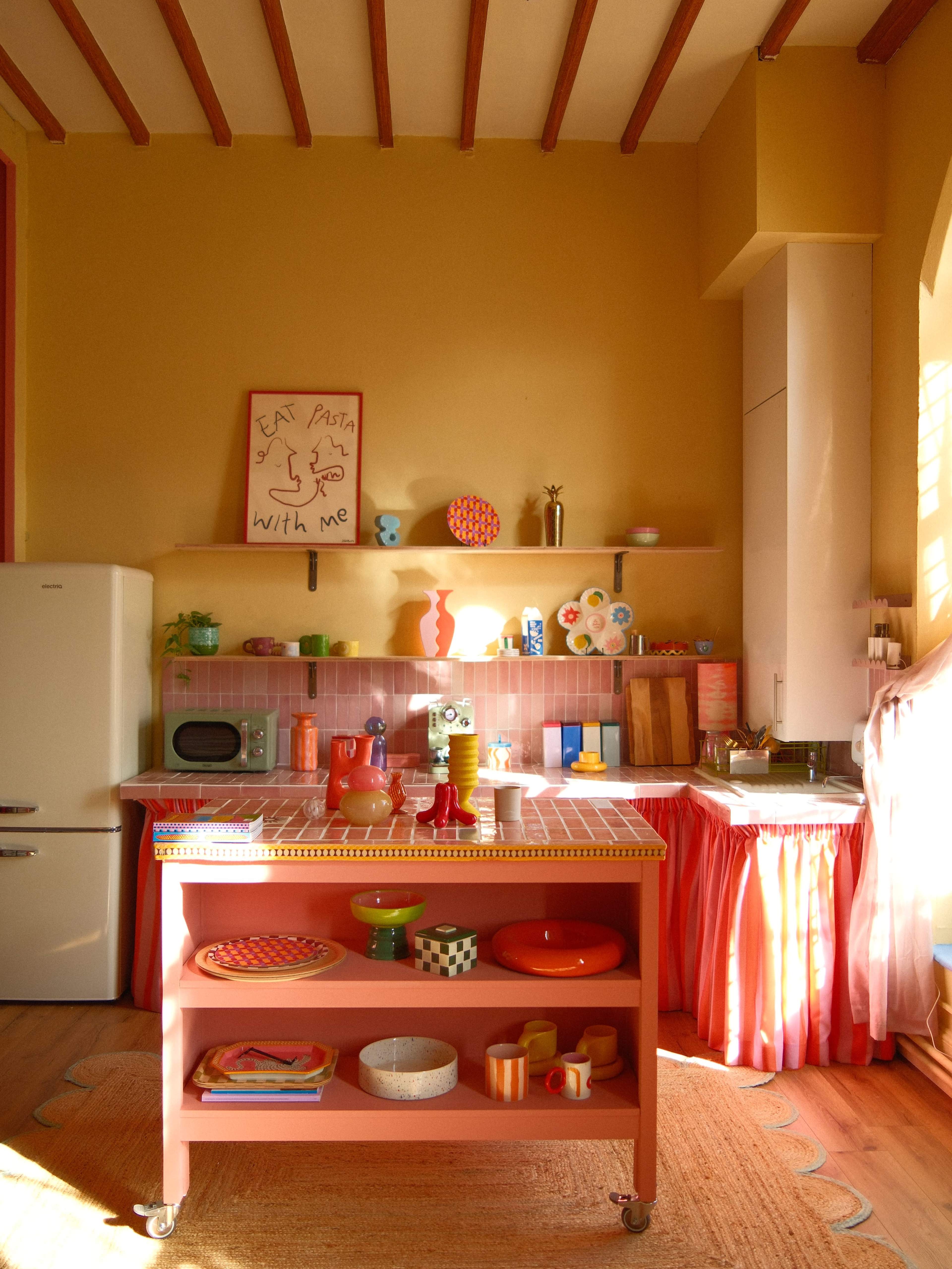 A colorful kitchen features a pink cart with various kitchenware, a retro refrigerator, and shelves filled with decorative items and appliances.