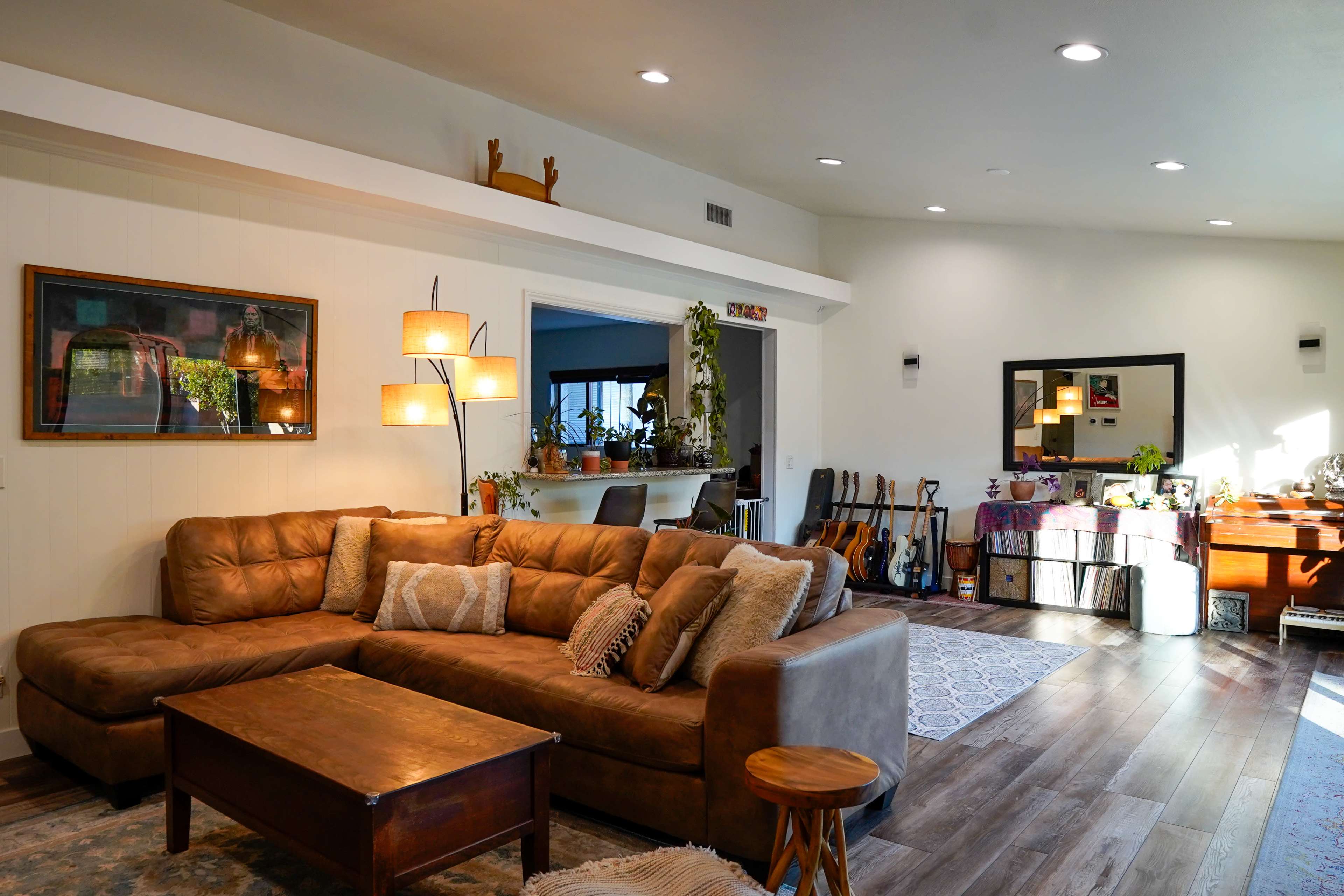 The image shows a well-lit living room featuring a large brown sectional sofa, a wooden coffee table, and various decor items, with an adjacent space that includes a bar area and shelves.