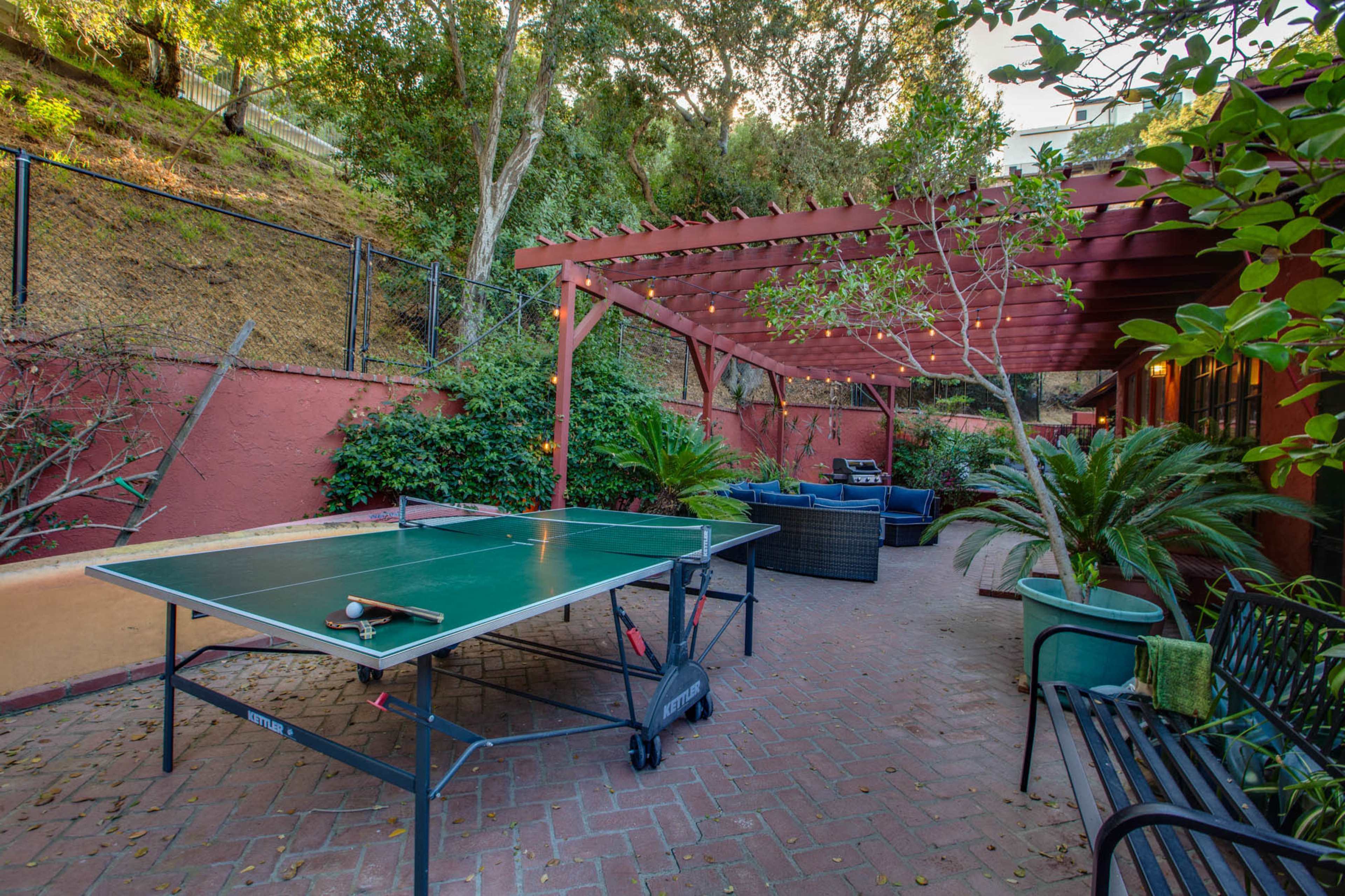The image shows a backyard patio area featuring a ping pong table surrounded by greenery and outdoor seating under a pergola.