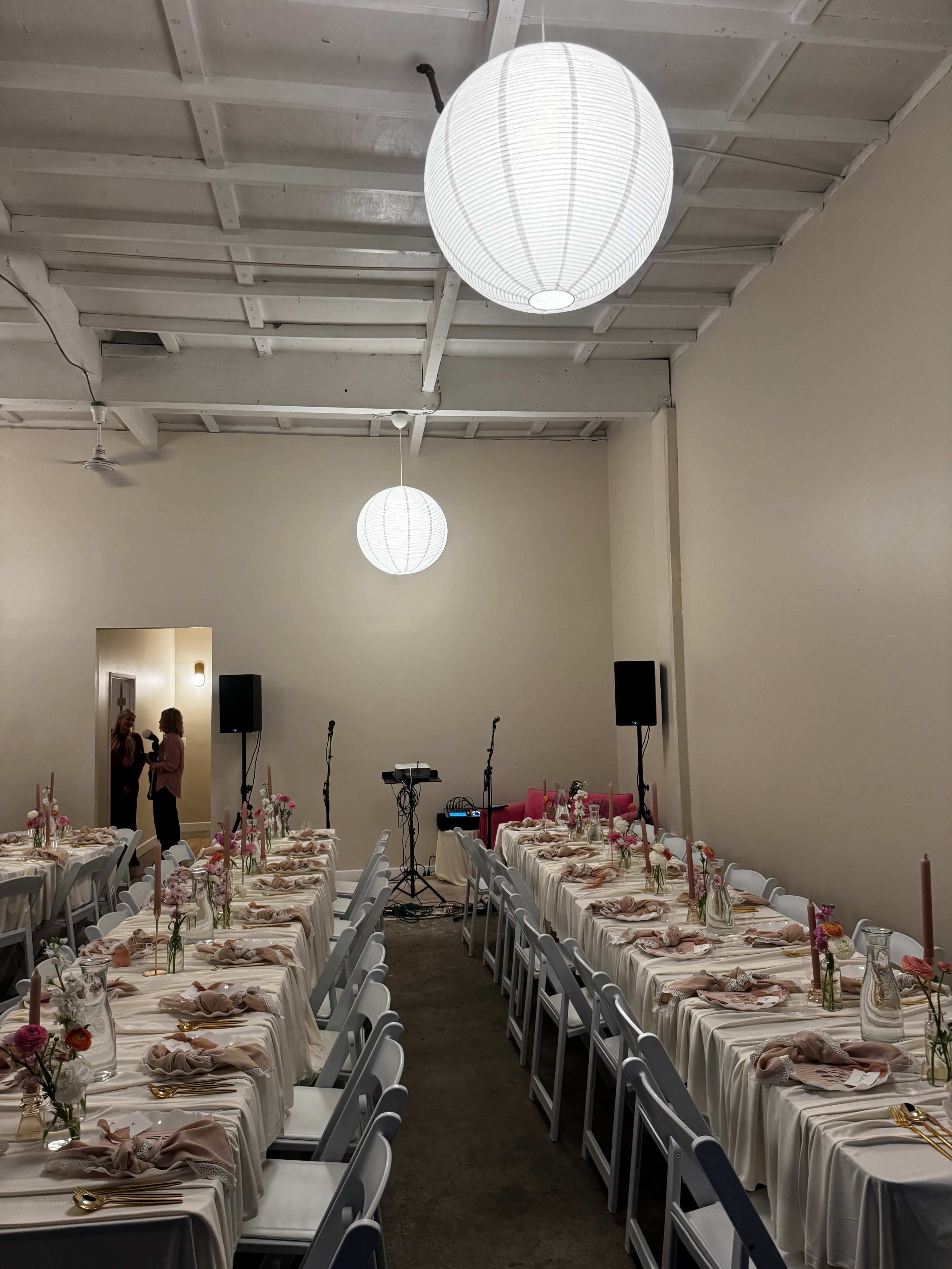 The image shows a decorated banquet hall with two long dining tables covered in white cloths, floral centerpieces, and pendant lights hanging from the ceiling.