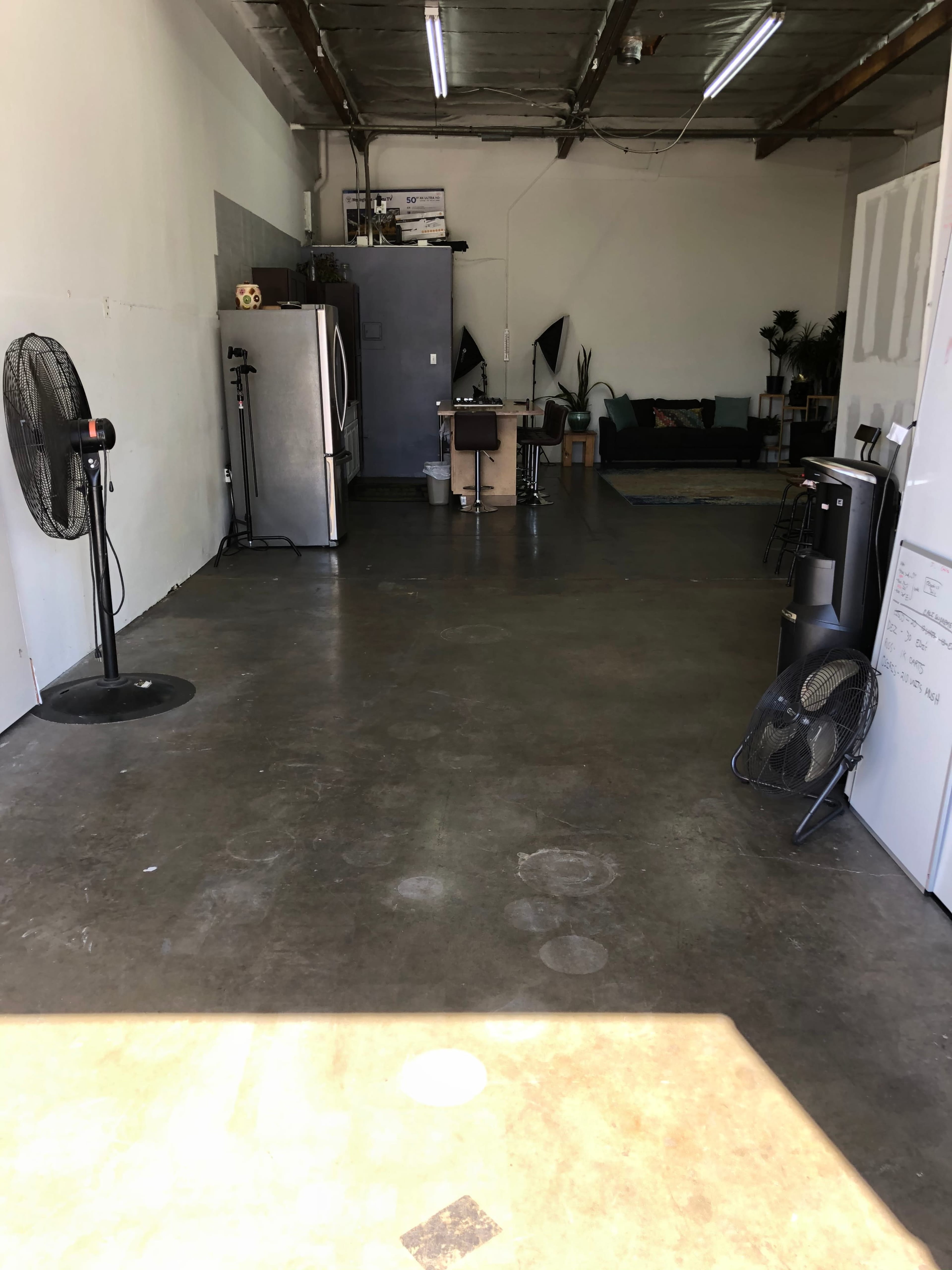 The image shows a spacious industrial-style room featuring appliances, a table with chairs, a sofa, and several fans.