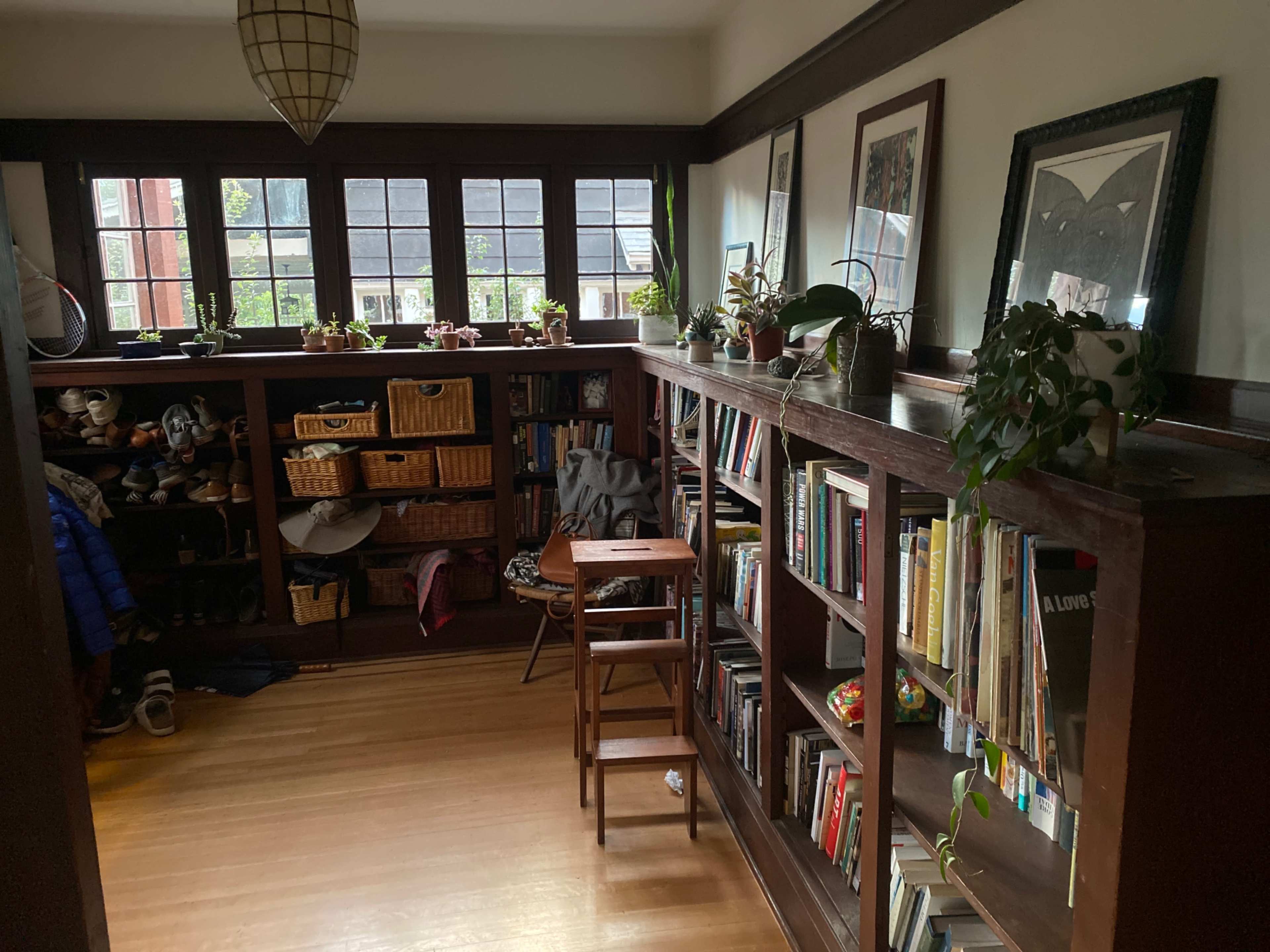 A small room with wooden shelves filled with books and decorative plants, along with a folding chair and storage baskets.