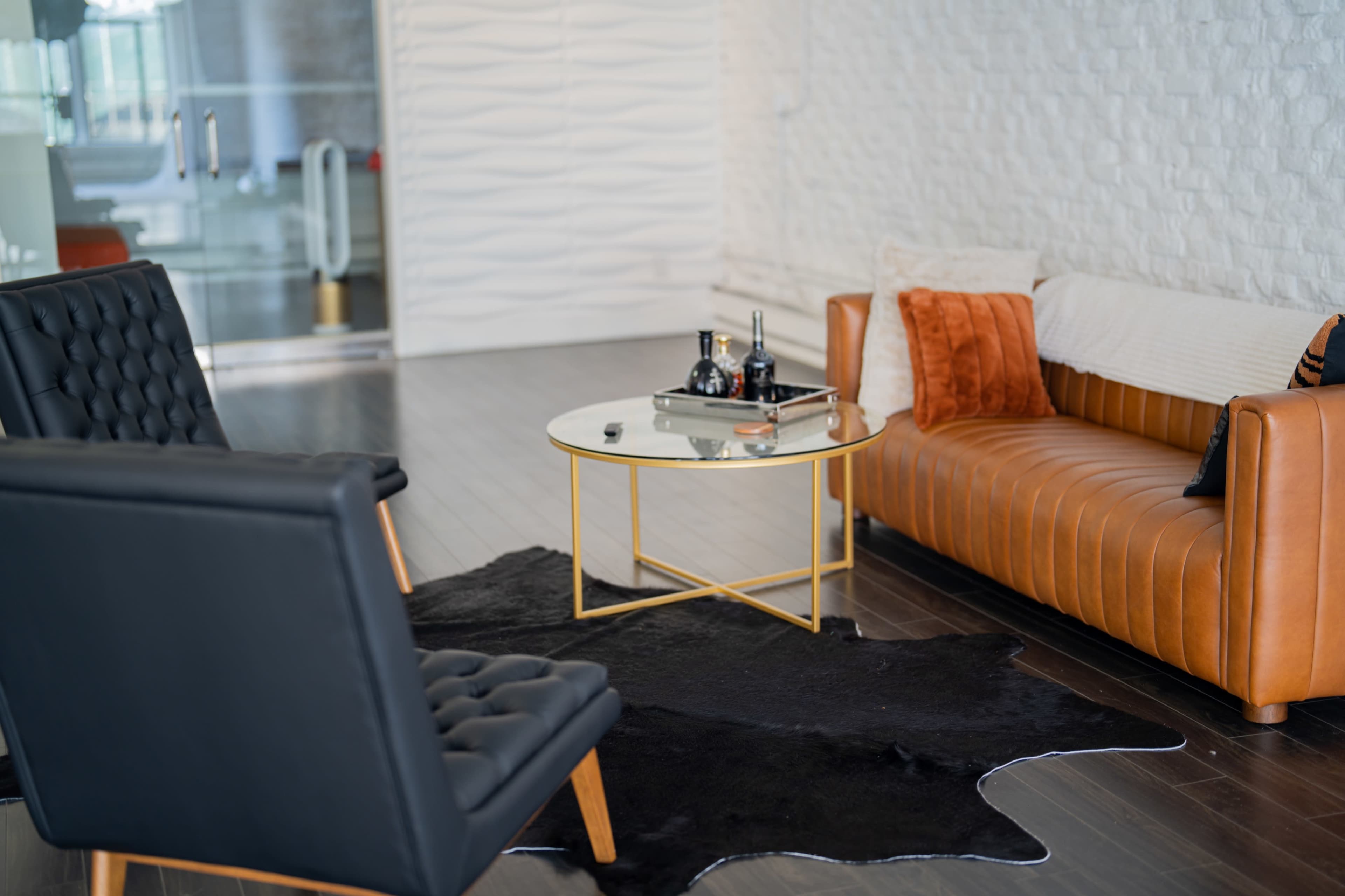 A modern living room with a brown leather sofa, two black lounge chairs, a round glass coffee table, and a cowhide rug on a dark wooden floor.