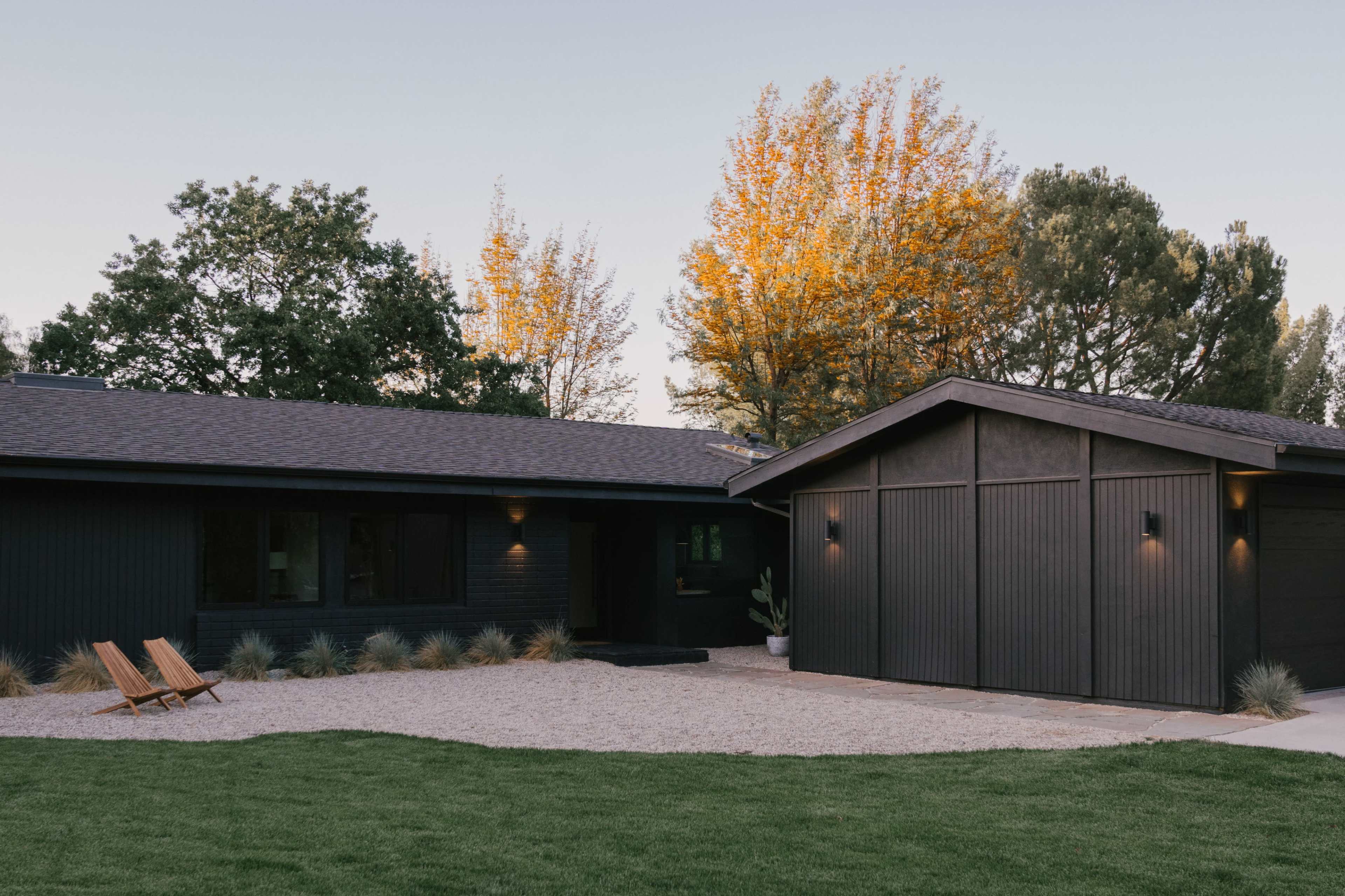 A modern black house with a sloped roof is surrounded by green grass and landscaped trees, featuring two wooden chairs on a gravel pathway.