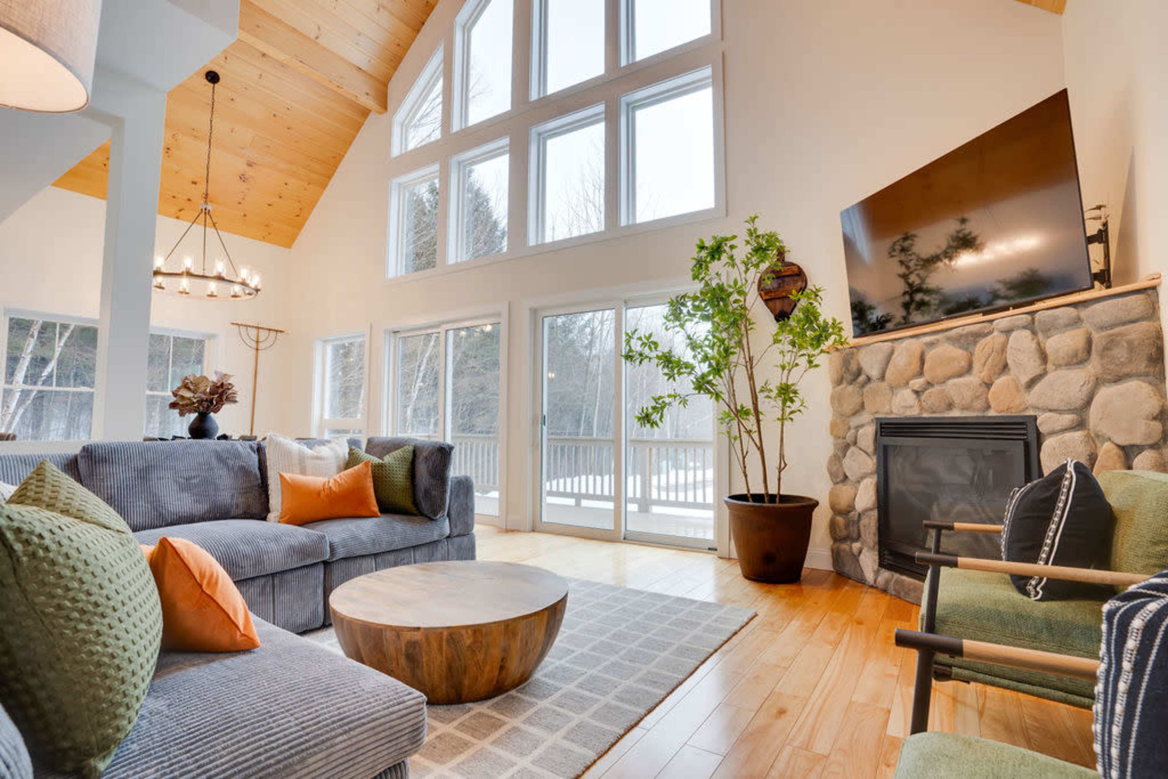 The image shows a modern living room with large windows, a stone fireplace, and a wooden ceiling, furnished with a gray sofa, a round wooden coffee table, and a green chair.