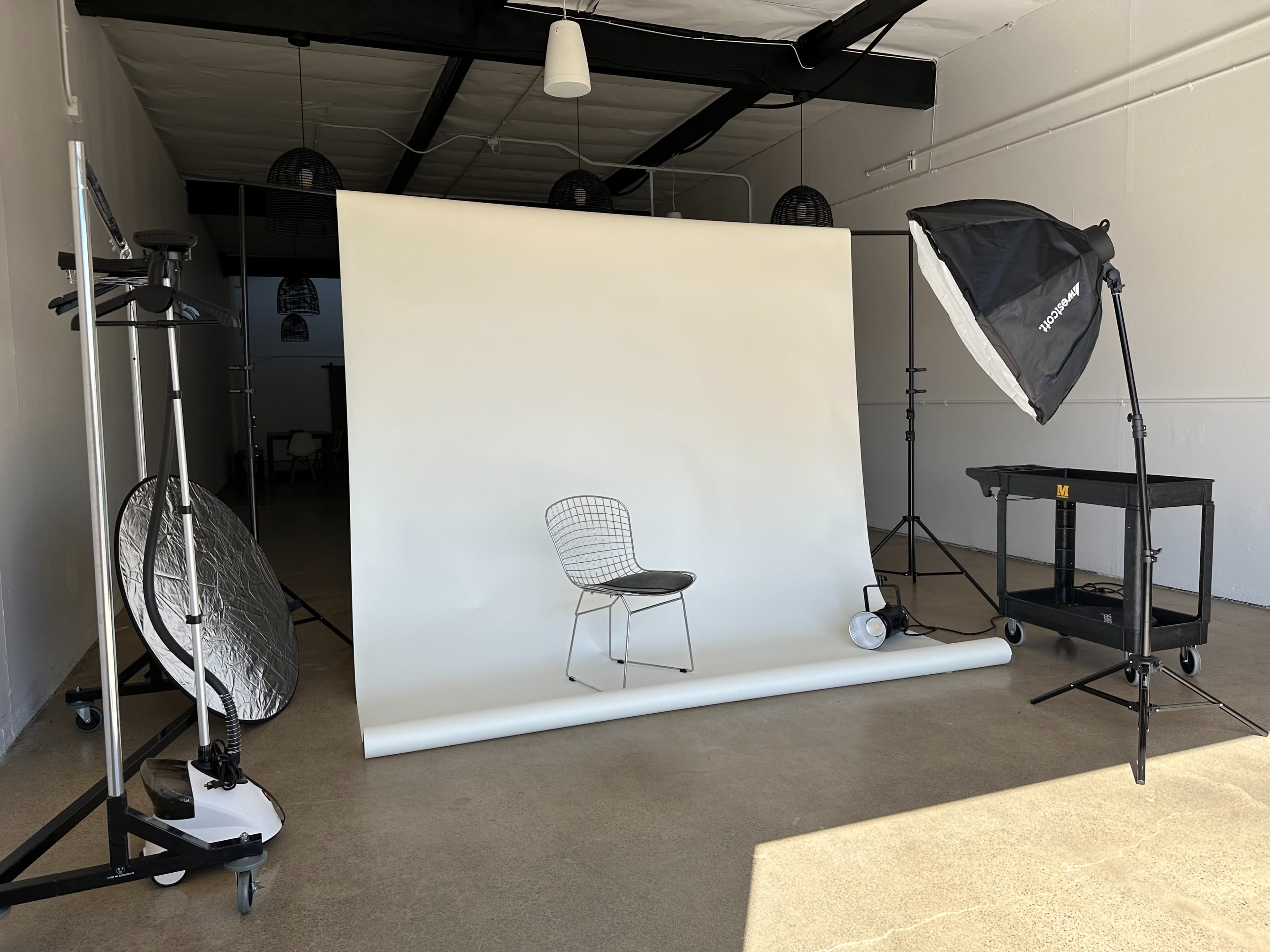 A photography studio features a white backdrop with a single chair in front, surrounded by lighting equipment and reflectors.