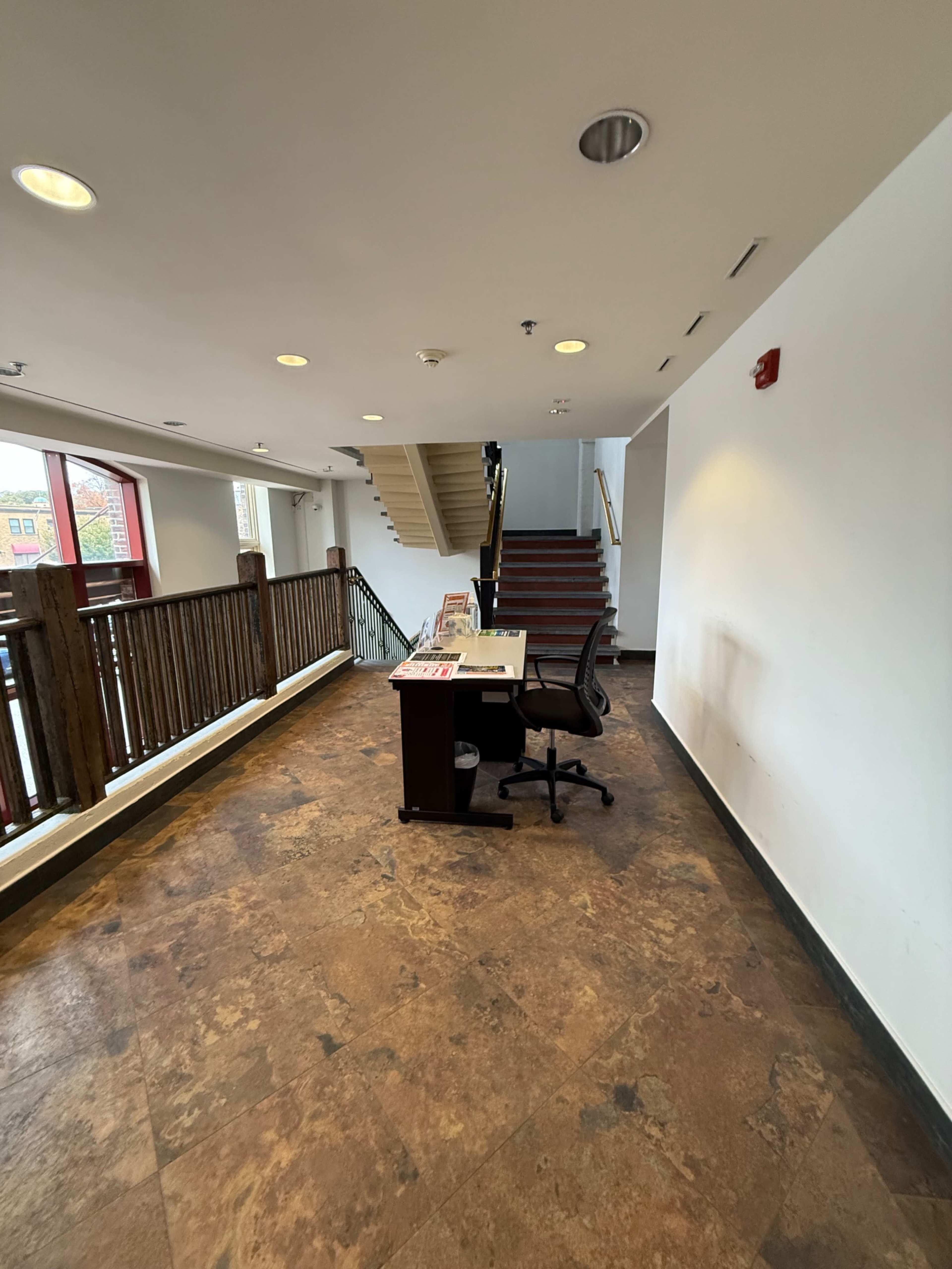 A small reception area features a desk with a chair, located beside a staircase and surrounded by a wooden railing.
