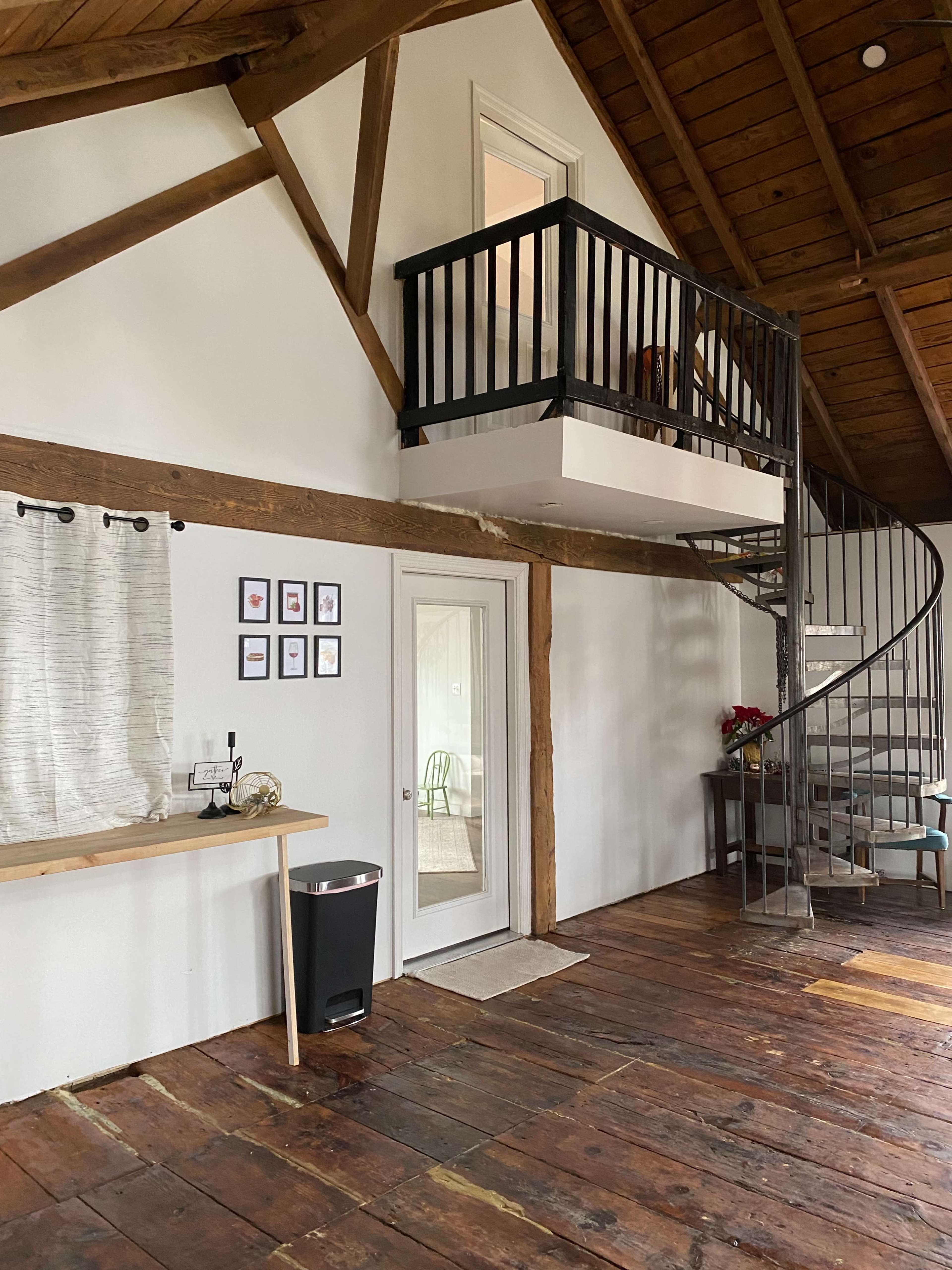 A spacious interior with a wooden floor, exposed beams, a spiral staircase to a loft area, and a small table displaying framed pictures.