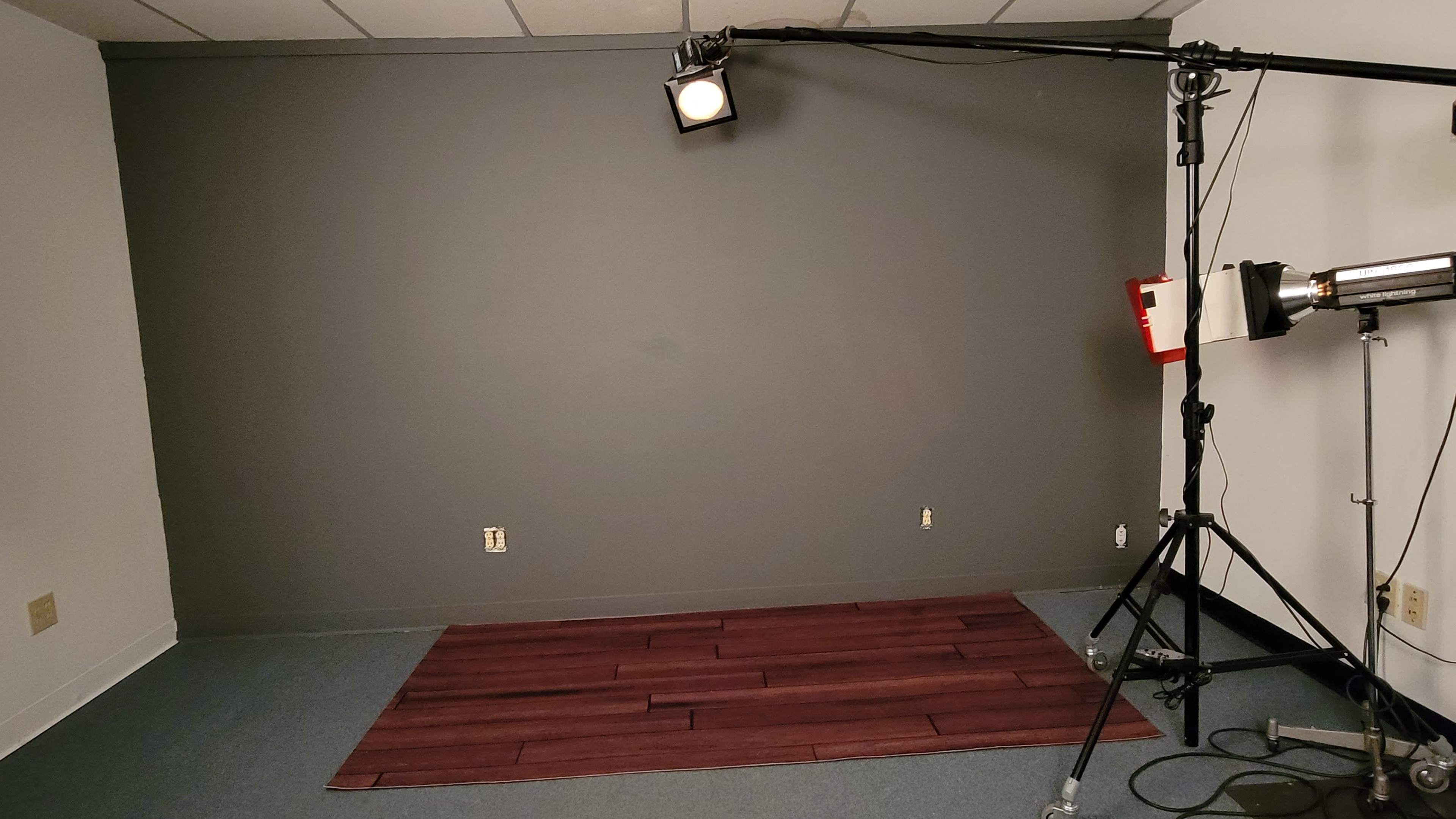 The image shows a sparse room with a gray wall, a spotlight mounted on a tripod, and a wooden mat on the floor.