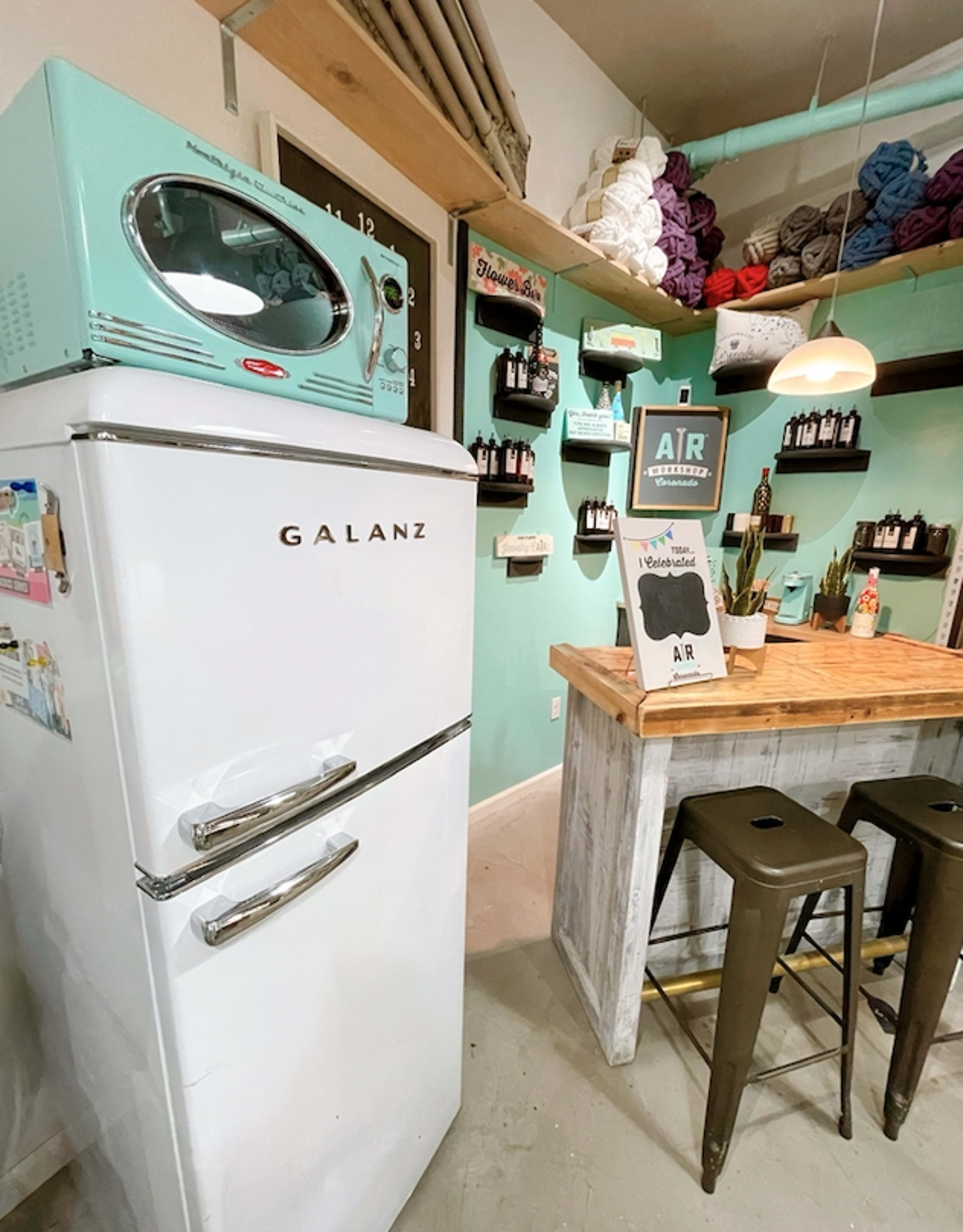 A retro-style white refrigerator next to a wooden table with metal stools in a brightly colored room adorned with shelves of yarn and craft supplies.