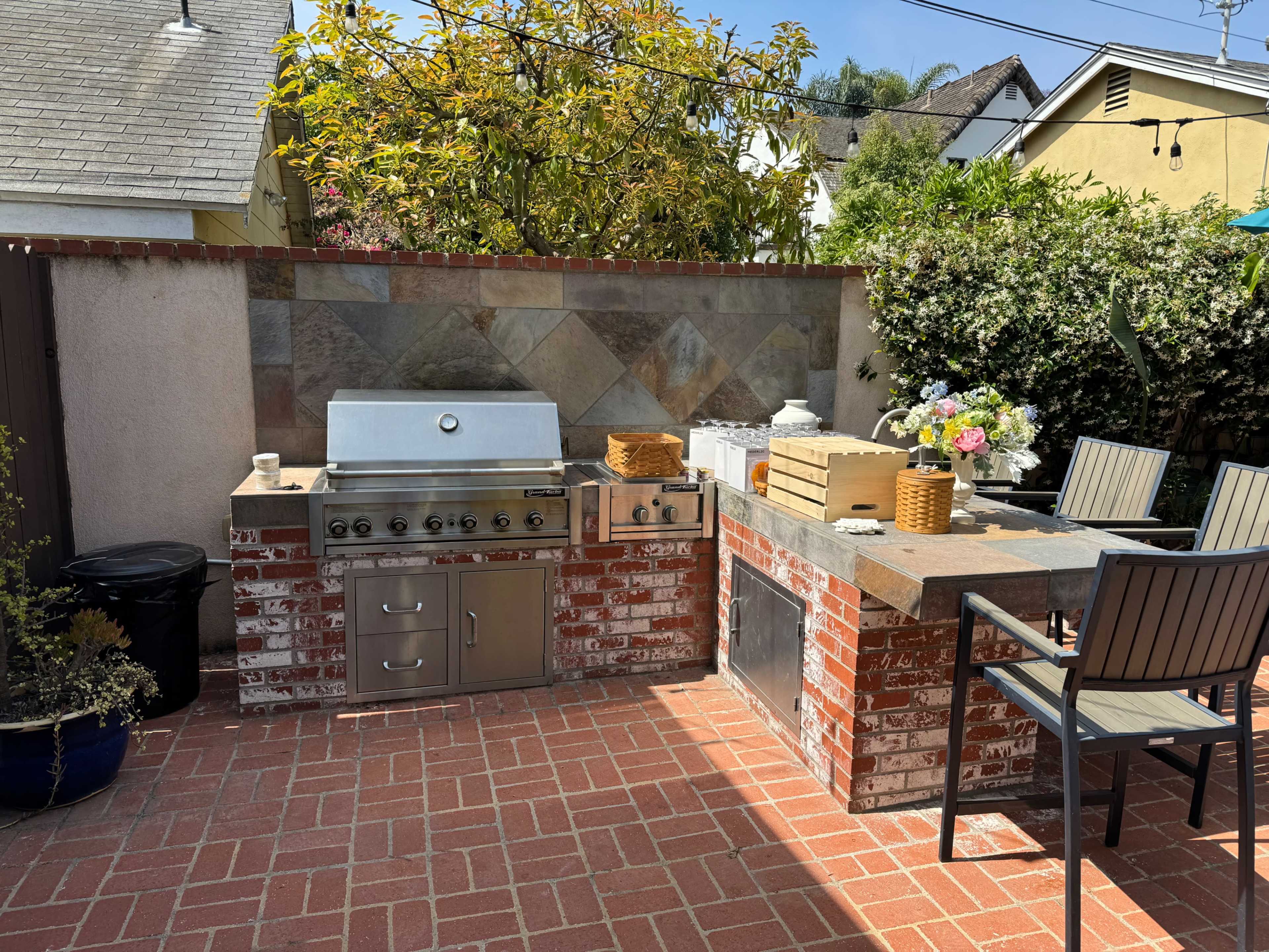 The image shows an outdoor kitchen area with a built-in grill, countertop, and seating surrounded by greenery.