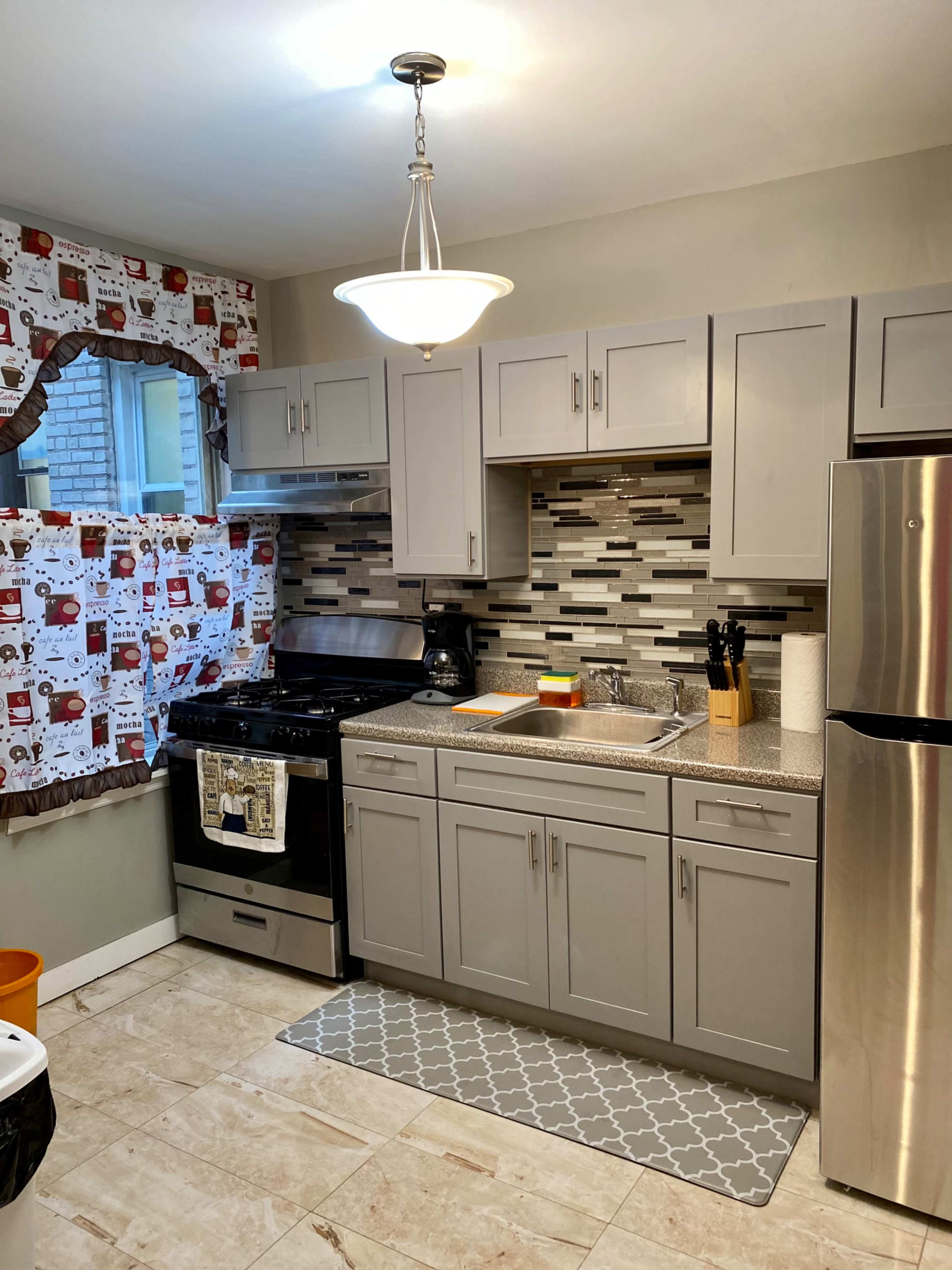 The kitchen features grey cabinets, a tiled backsplash, a stainless steel refrigerator, and a gas stove with a colorful cutting board on the countertop.