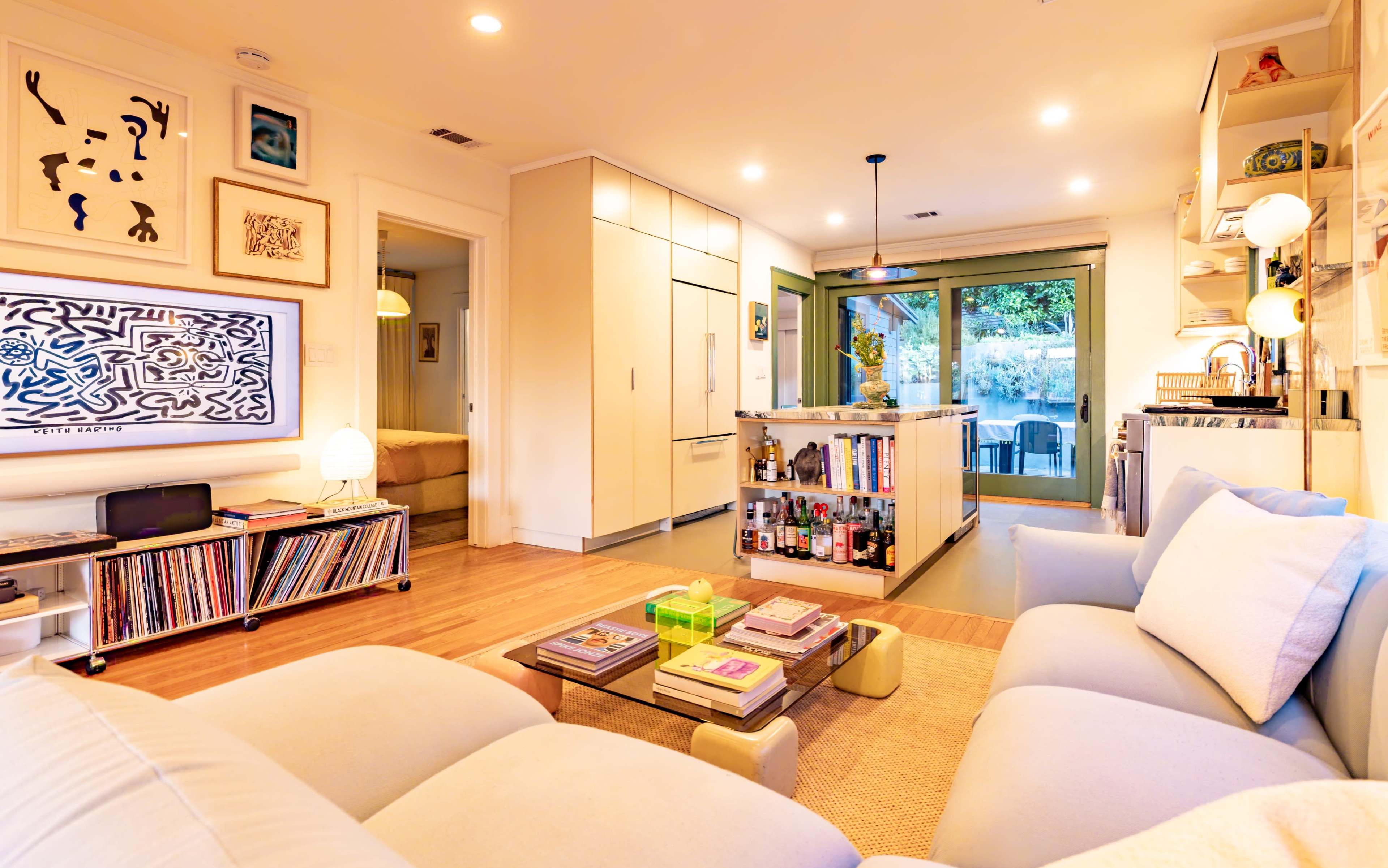 The image shows a contemporary living room with a beige sectional sofa, a coffee table displaying magazines, and an open layout connecting to a kitchen and sliding glass doors leading to an outdoor space.