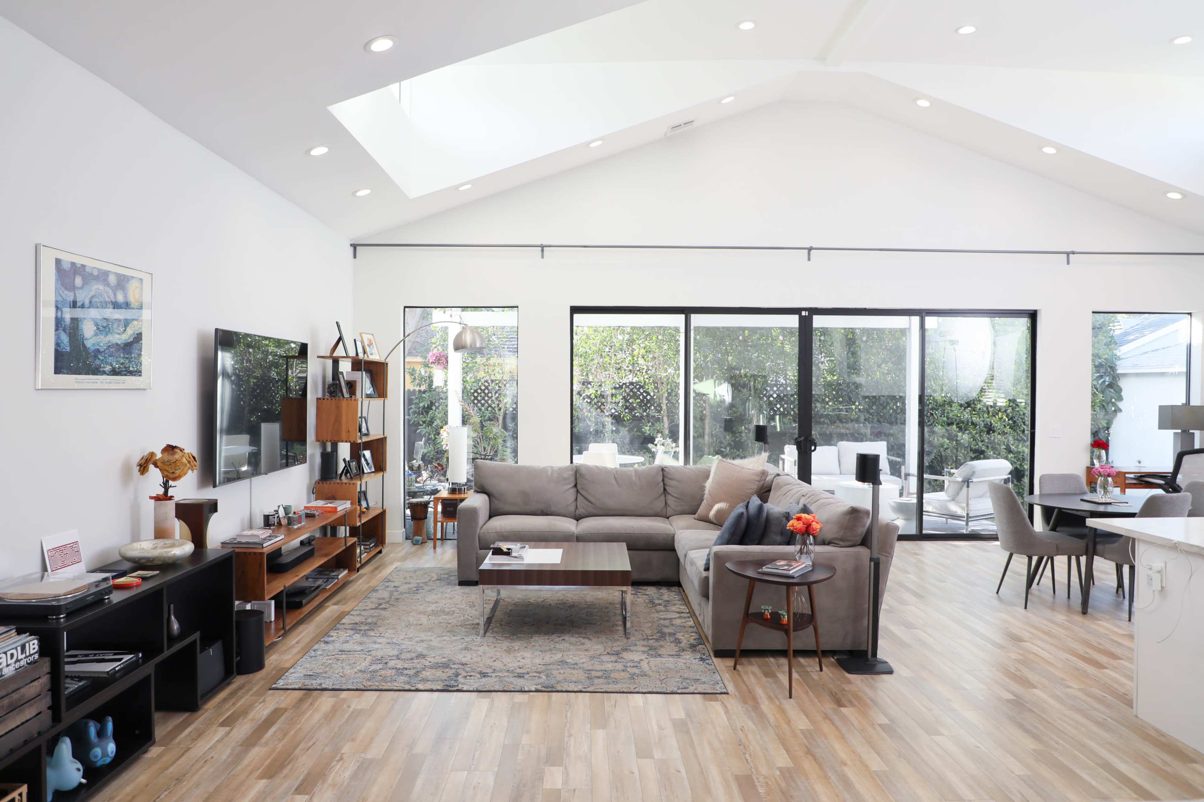 A modern living room features a gray sectional sofa, wooden furniture, a large TV, and large windows looking out to a patio area.