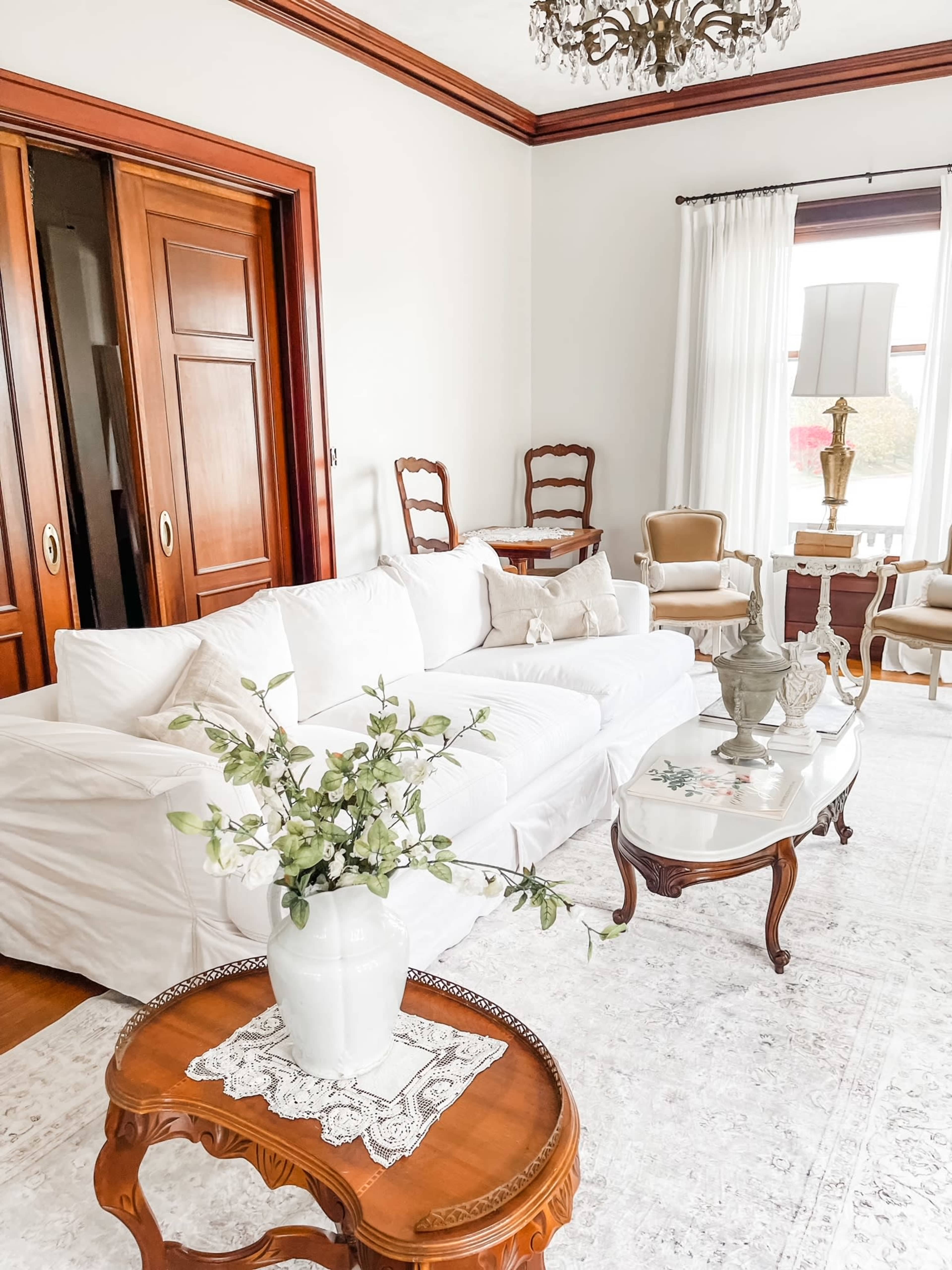 A cozy living room featuring a white sofa, a wooden coffee table with a lace doily, and vintage chairs under a chandelier.