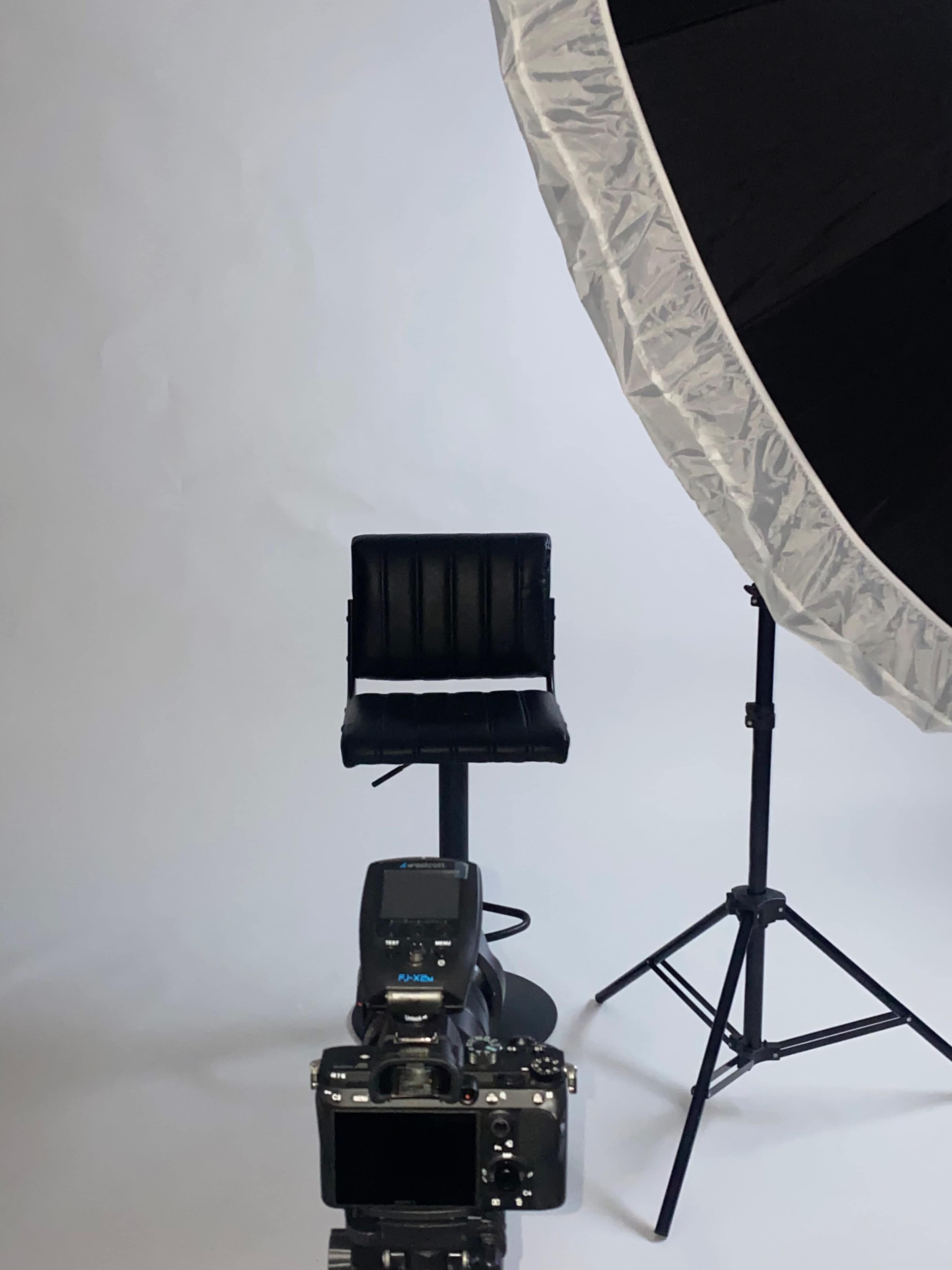 A black stool is positioned in front of a camera and a large softbox light against a plain backdrop.