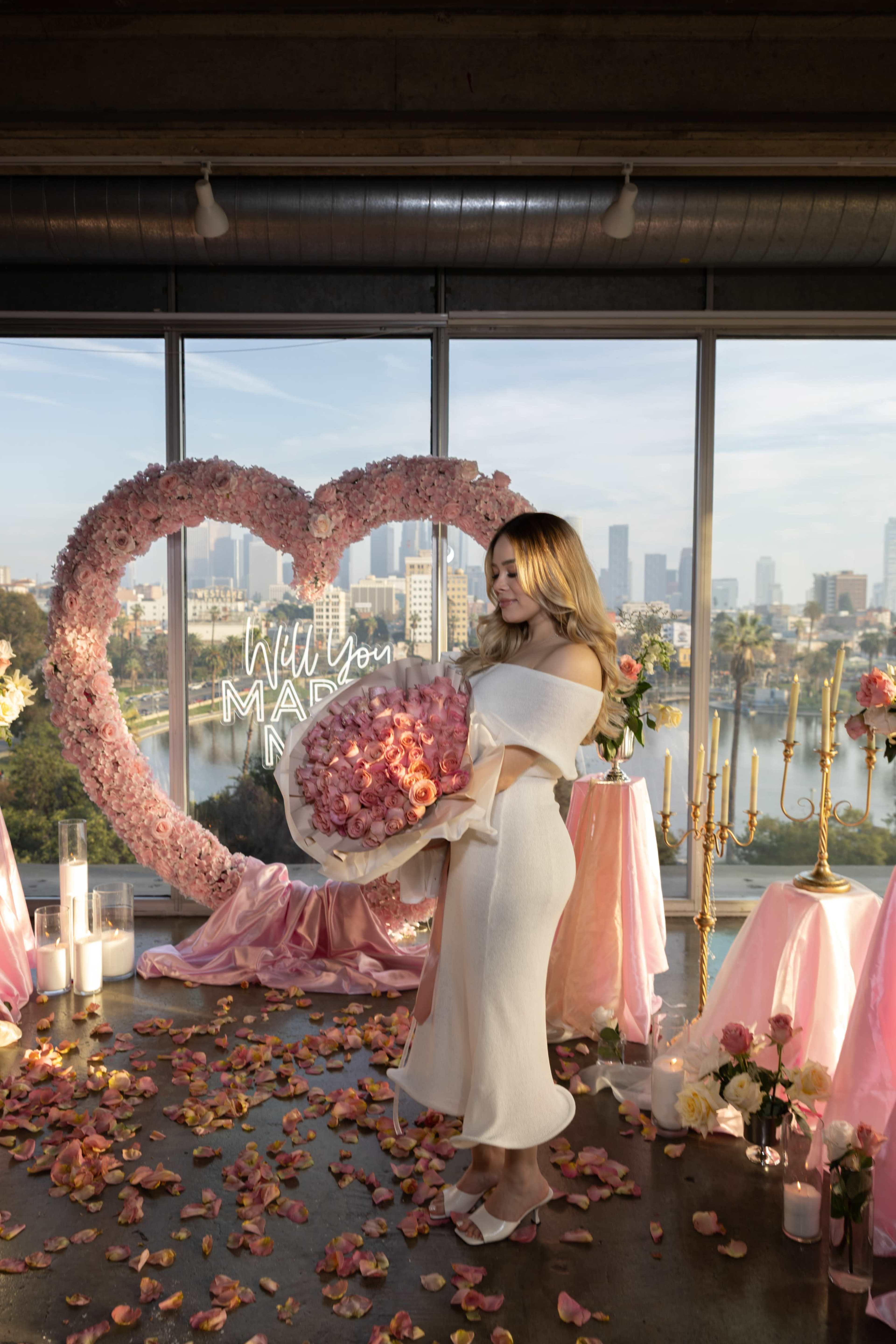 A woman in a white dress holds a large bouquet of pink roses in front of a heart-shaped floral display and a city skyline.
