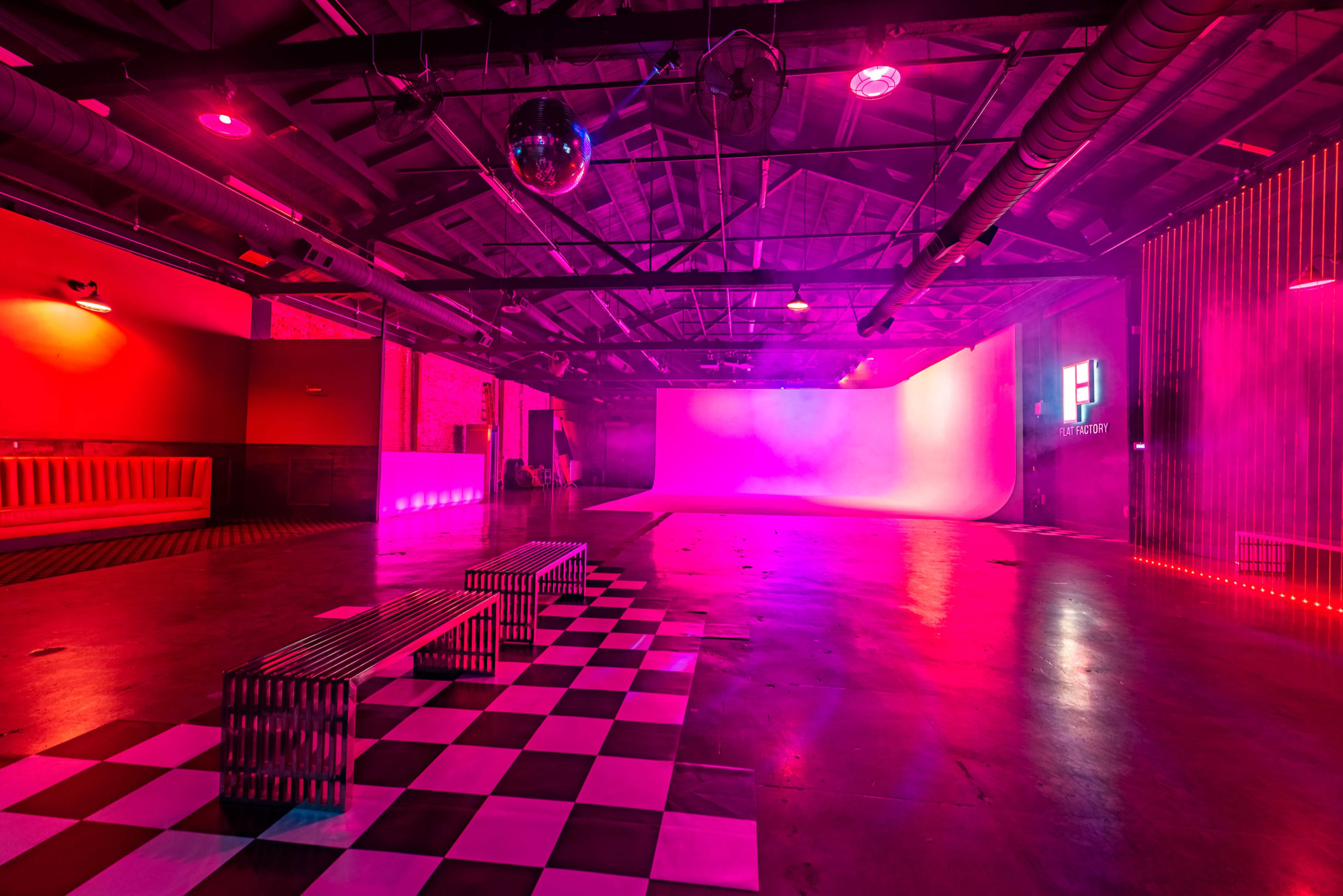 The image shows an empty industrial space with a checkered floor, pink lighting, and a white backdrop at one end.