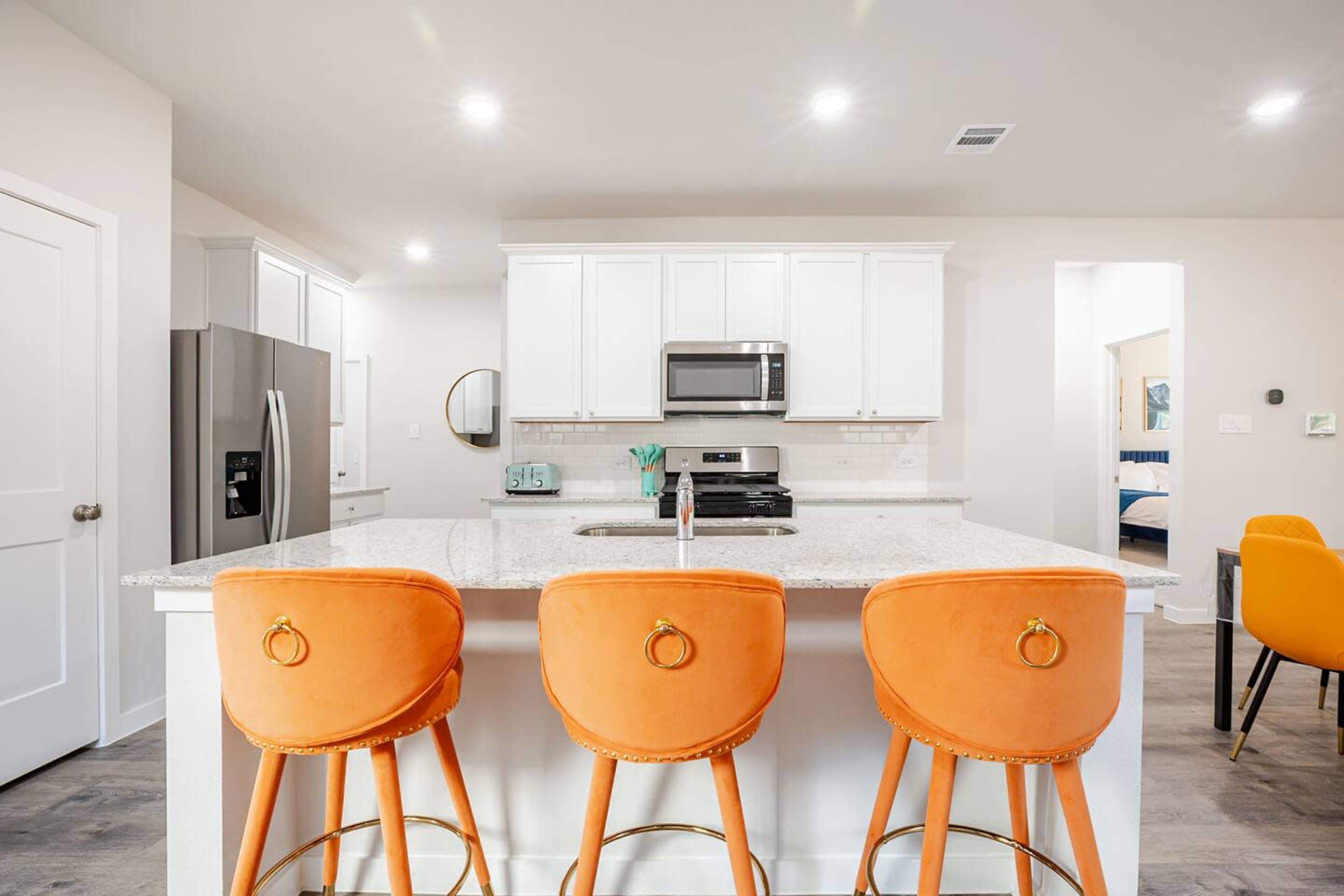 The image depicts a modern kitchen featuring a center island with three orange barstools, stainless steel appliances, and white cabinetry.
