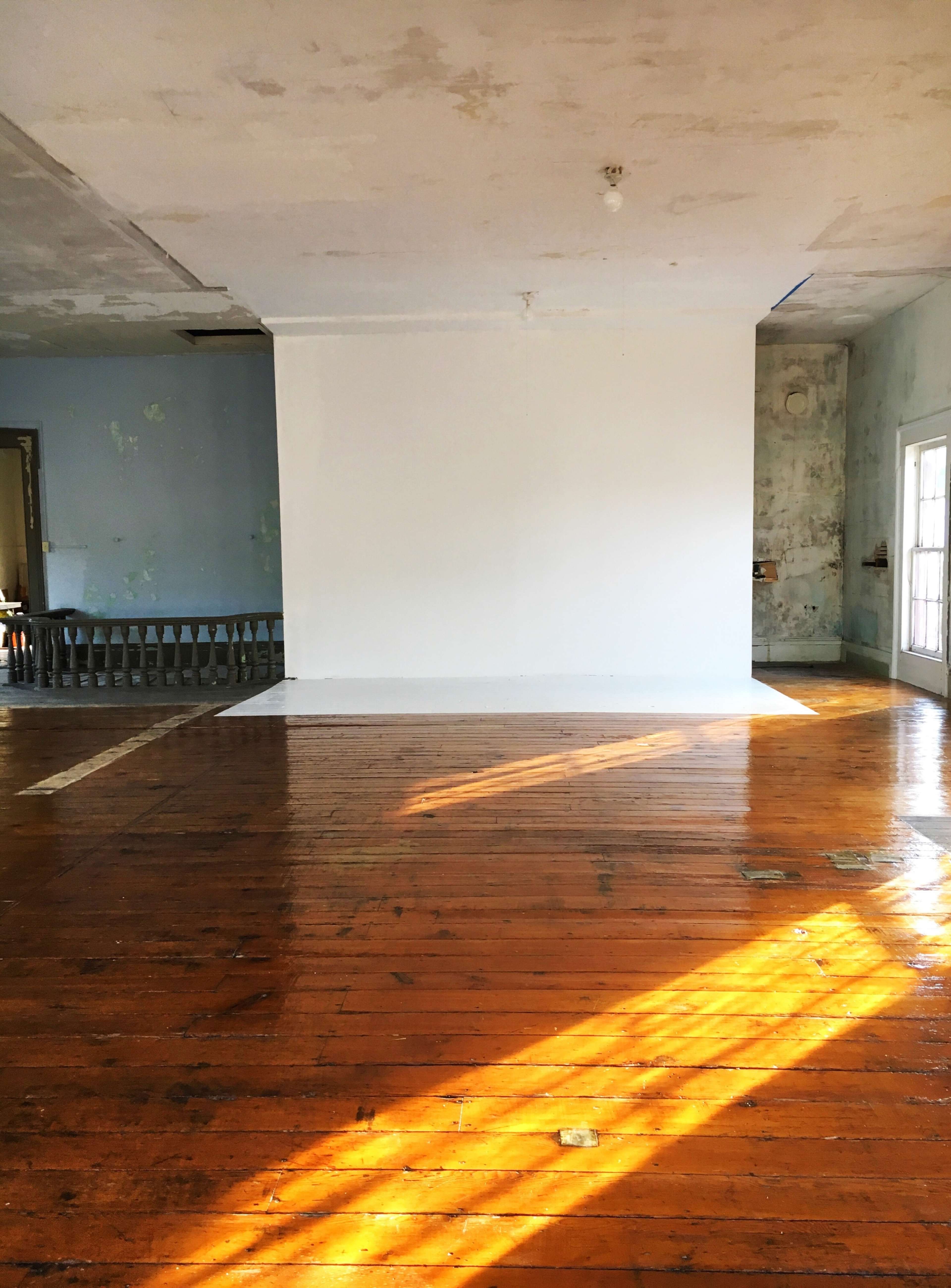 An empty room with wooden floors, a white wall at the center, and sunlight streaming through a window.