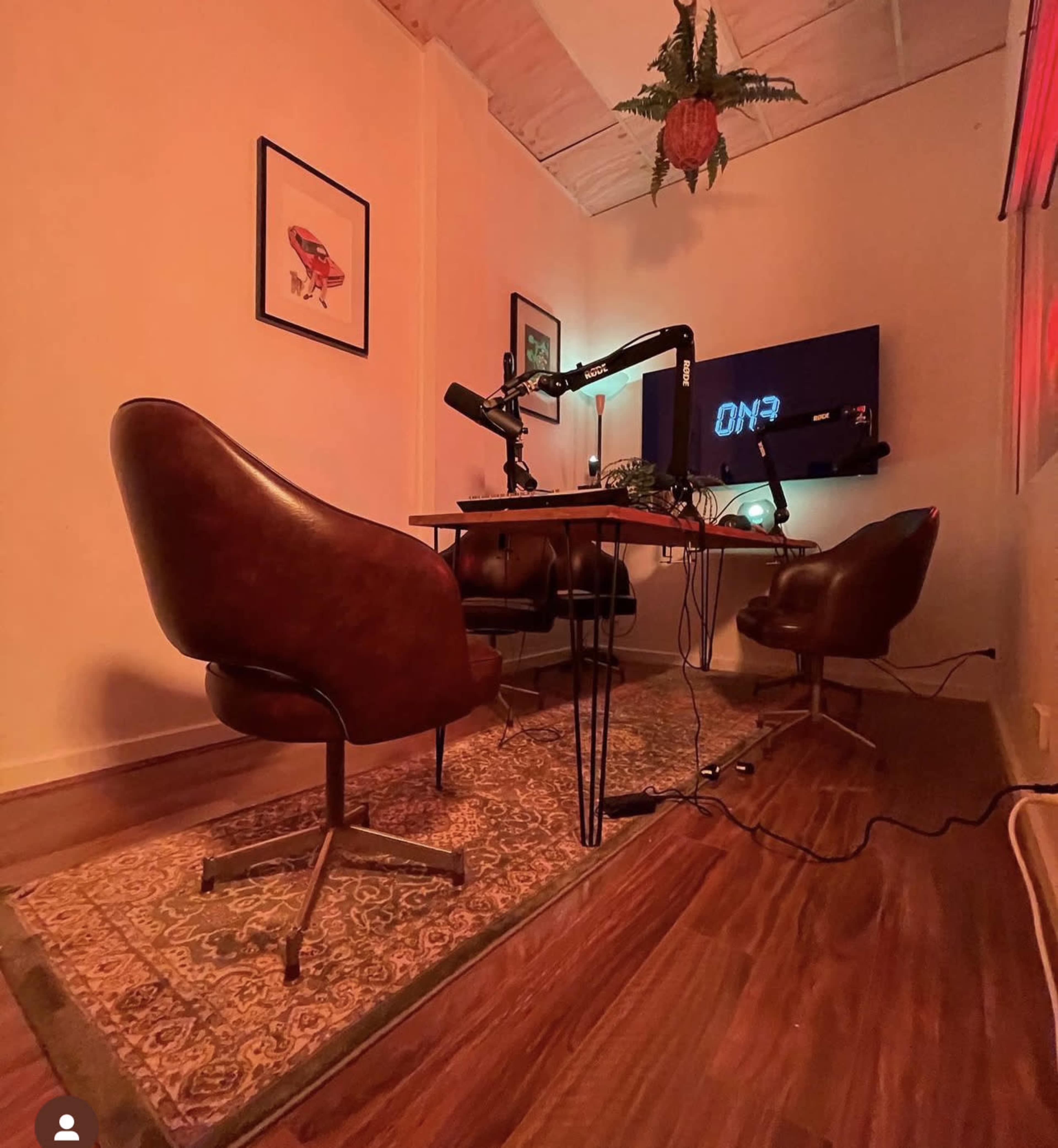 A small podcast studio featuring two brown chairs, a microphone setup on a table, and a screen displaying "ON3" on the wall.