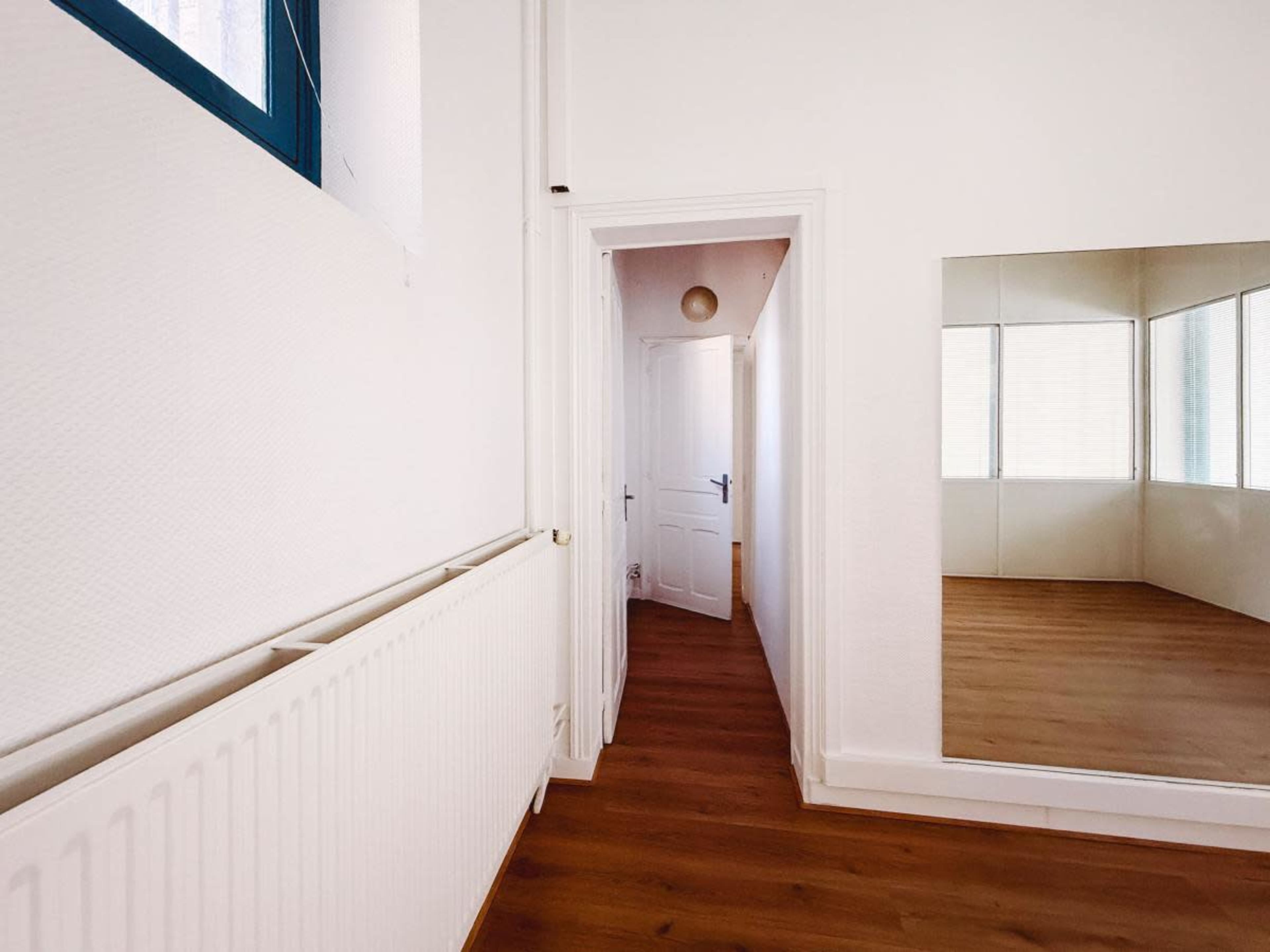 A hallway with a white wall, wooden floor, and a large mirror reflects an adjacent room.