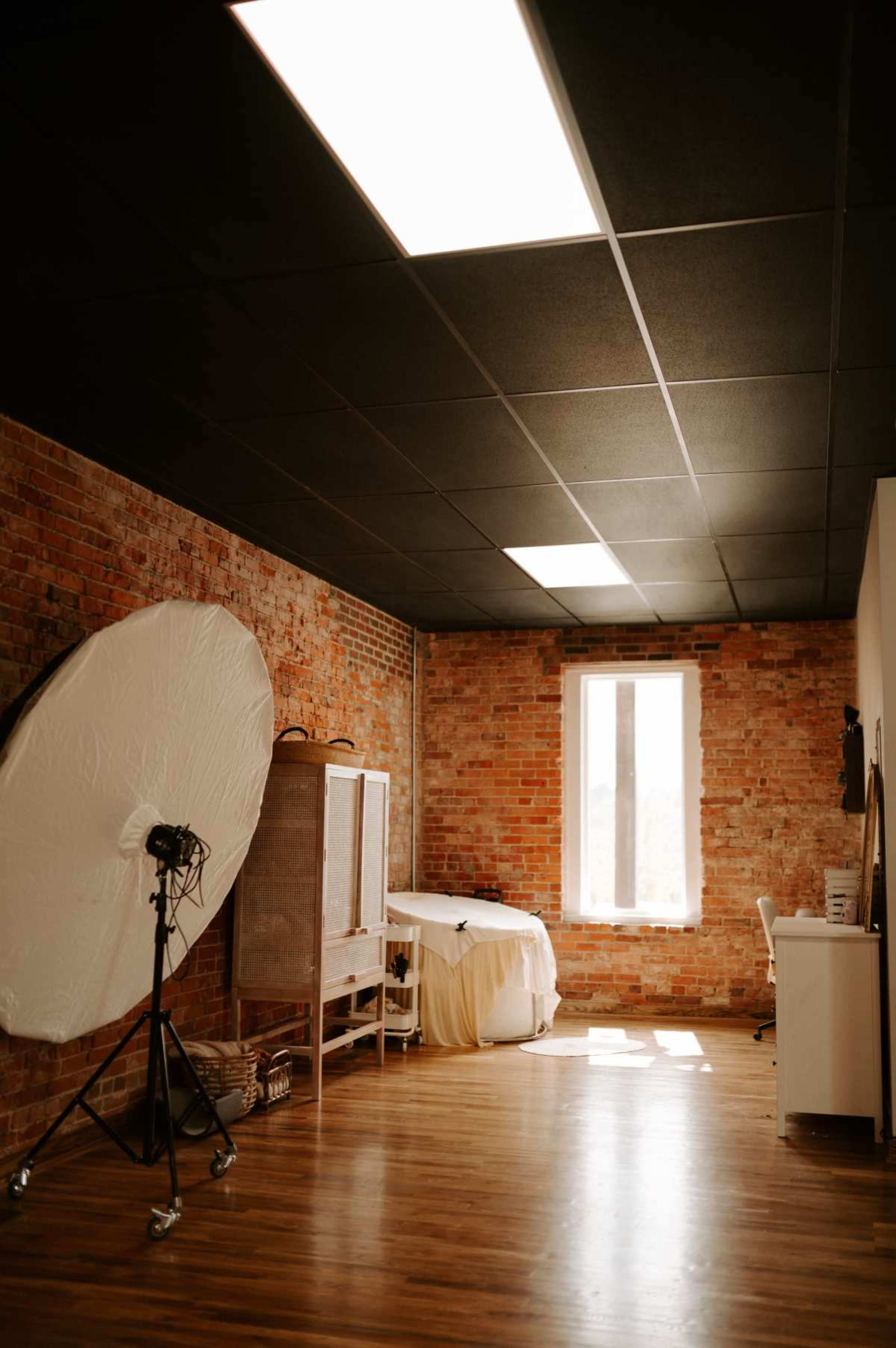 A spacious room with exposed brick walls, a large softbox light on a stand, a white curtain, and a window allowing natural light to enter.