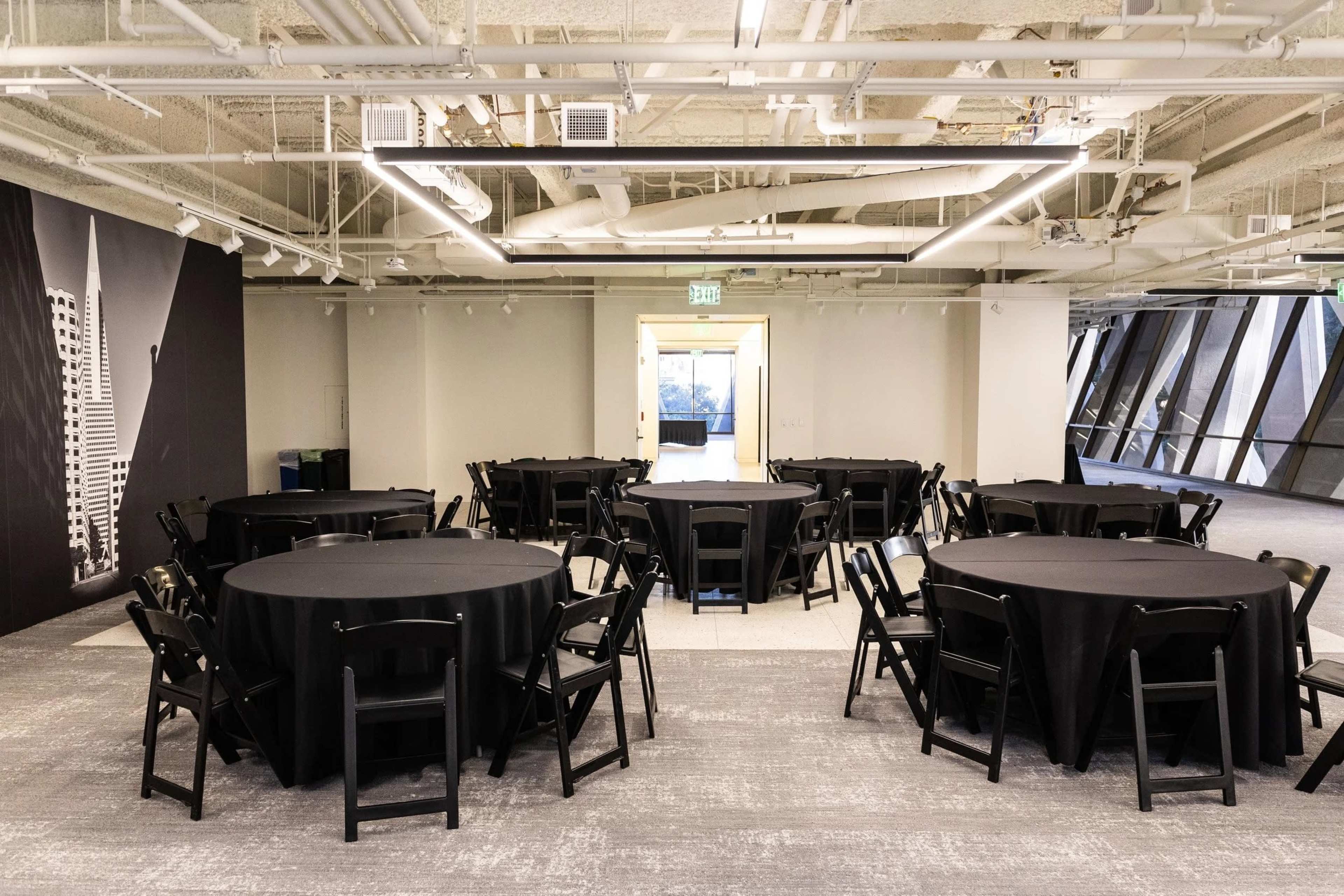 The image shows a large, empty event space arranged with round tables covered in black tablecloths and black chairs, with a door leading to another area in the background.
