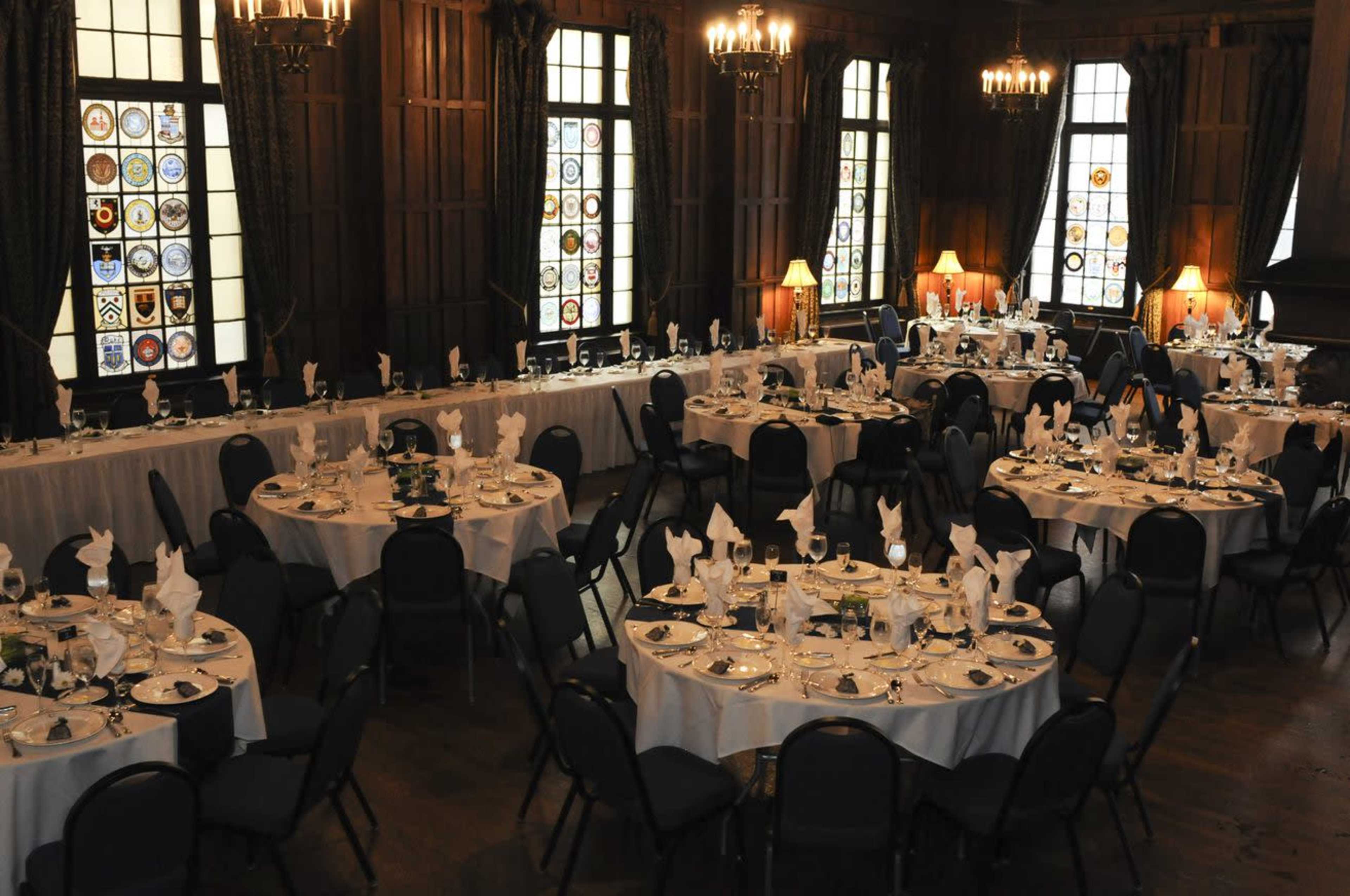 A formal dining hall set up with round tables covered with white tablecloths and arranged with place settings, surrounded by wooden walls and large stained glass windows.