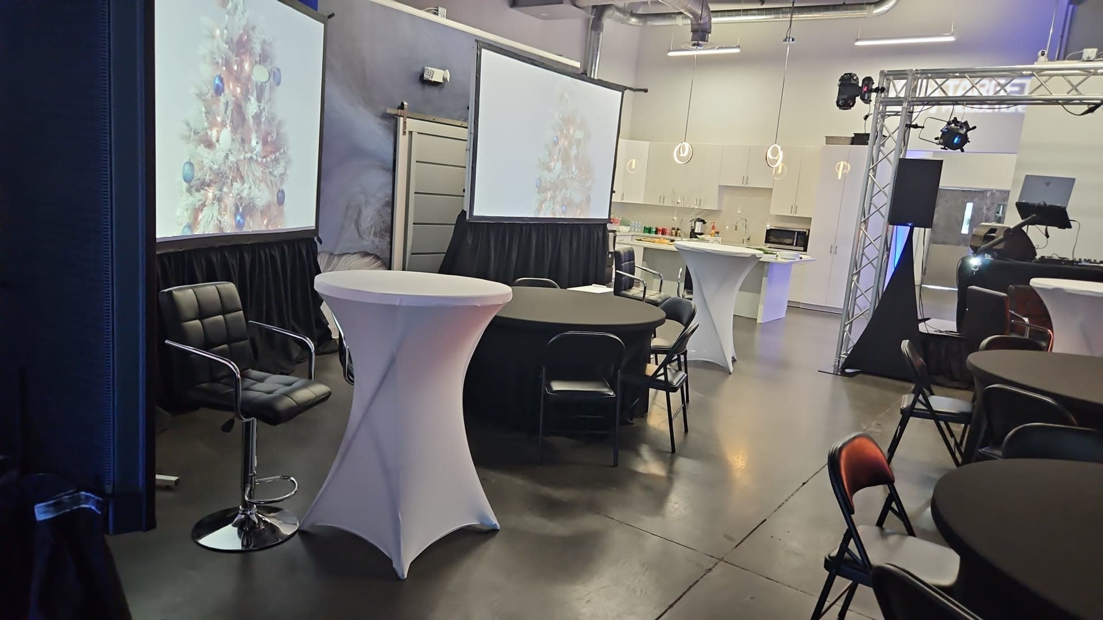 The image shows an event space furnished with several round tables, chairs, and two large screens displaying festive imagery, along with a bar area in the background.