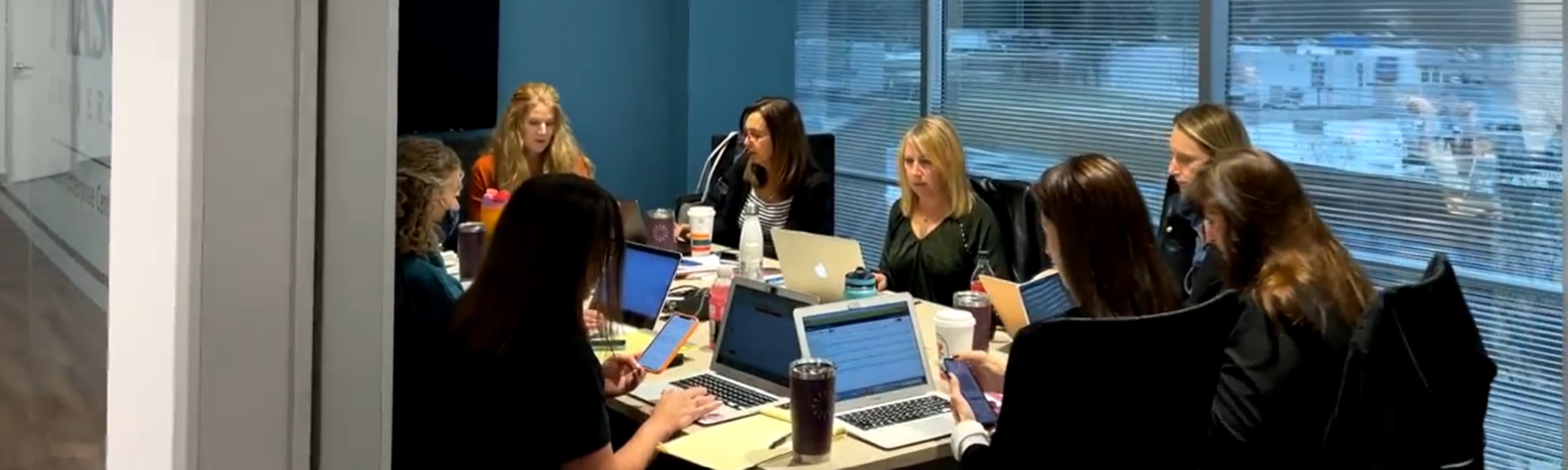 A group of eight women is sitting around a conference table, working on laptops and discussing materials in a modern office setting.