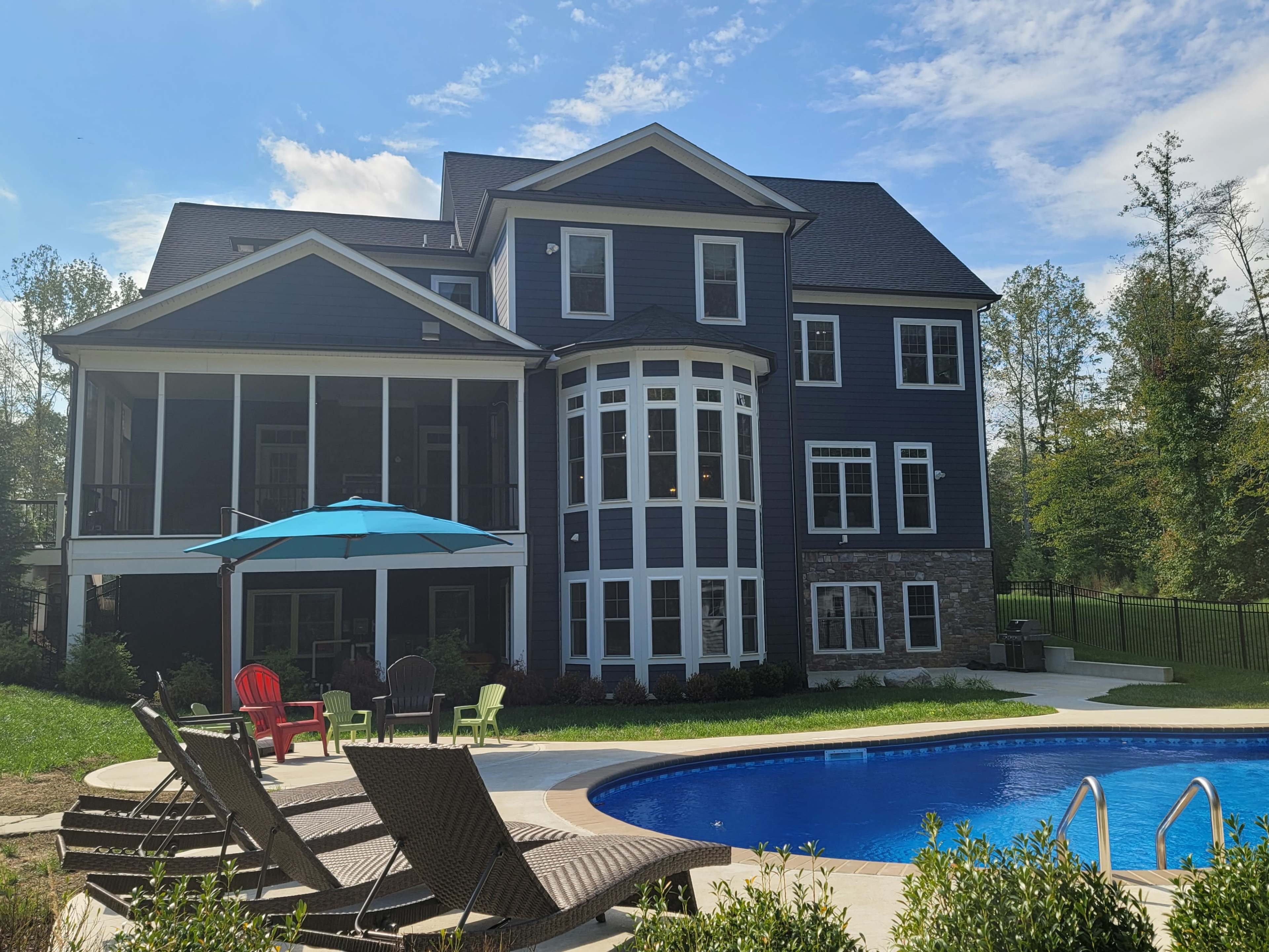 A large, modern house with a stone and dark siding exterior, a swimming pool in the foreground, and a patio with lounge chairs and an umbrella.
