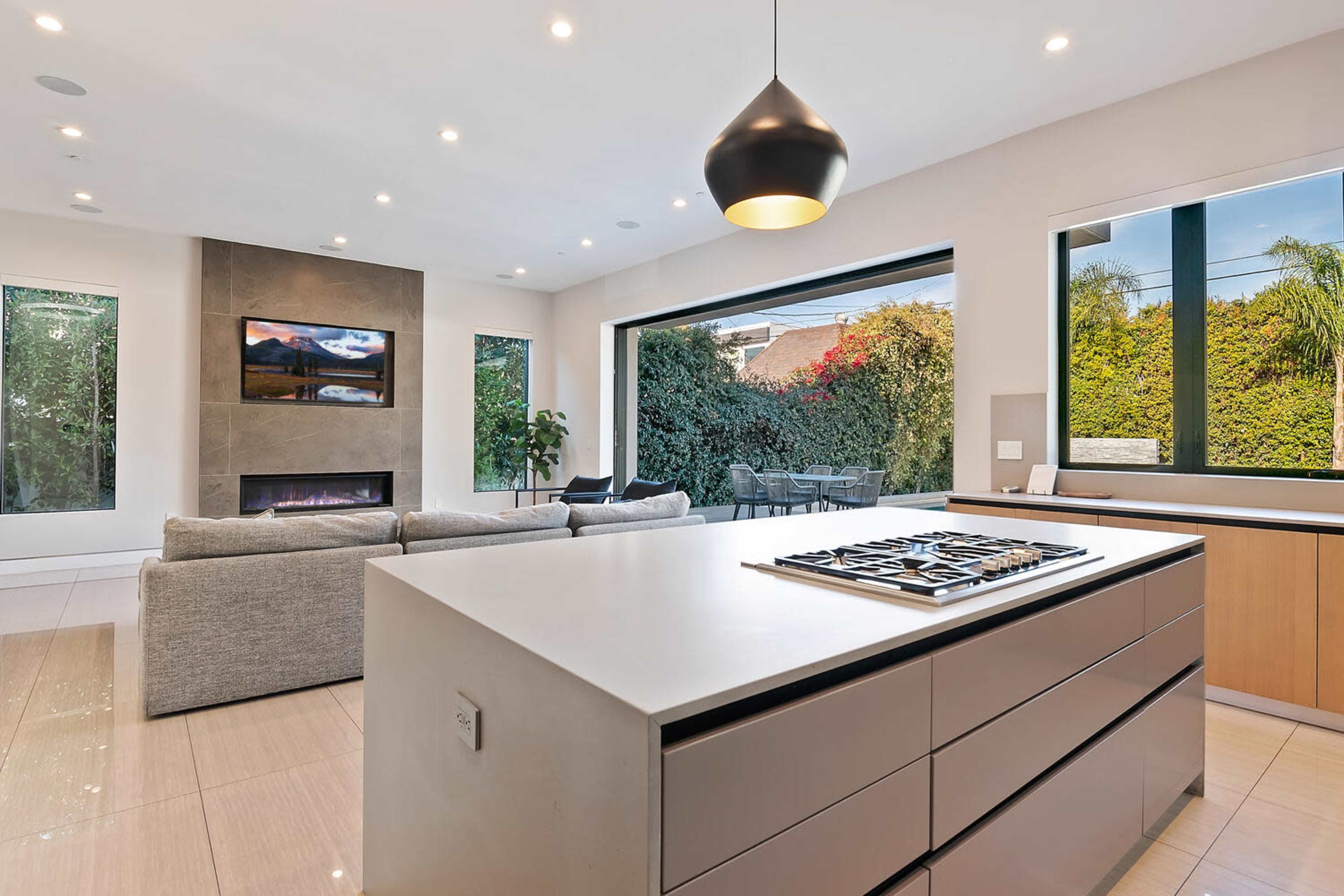 A modern kitchen features an island with a stovetop, open to a living area with a large window overlooking a landscaped outdoor space.