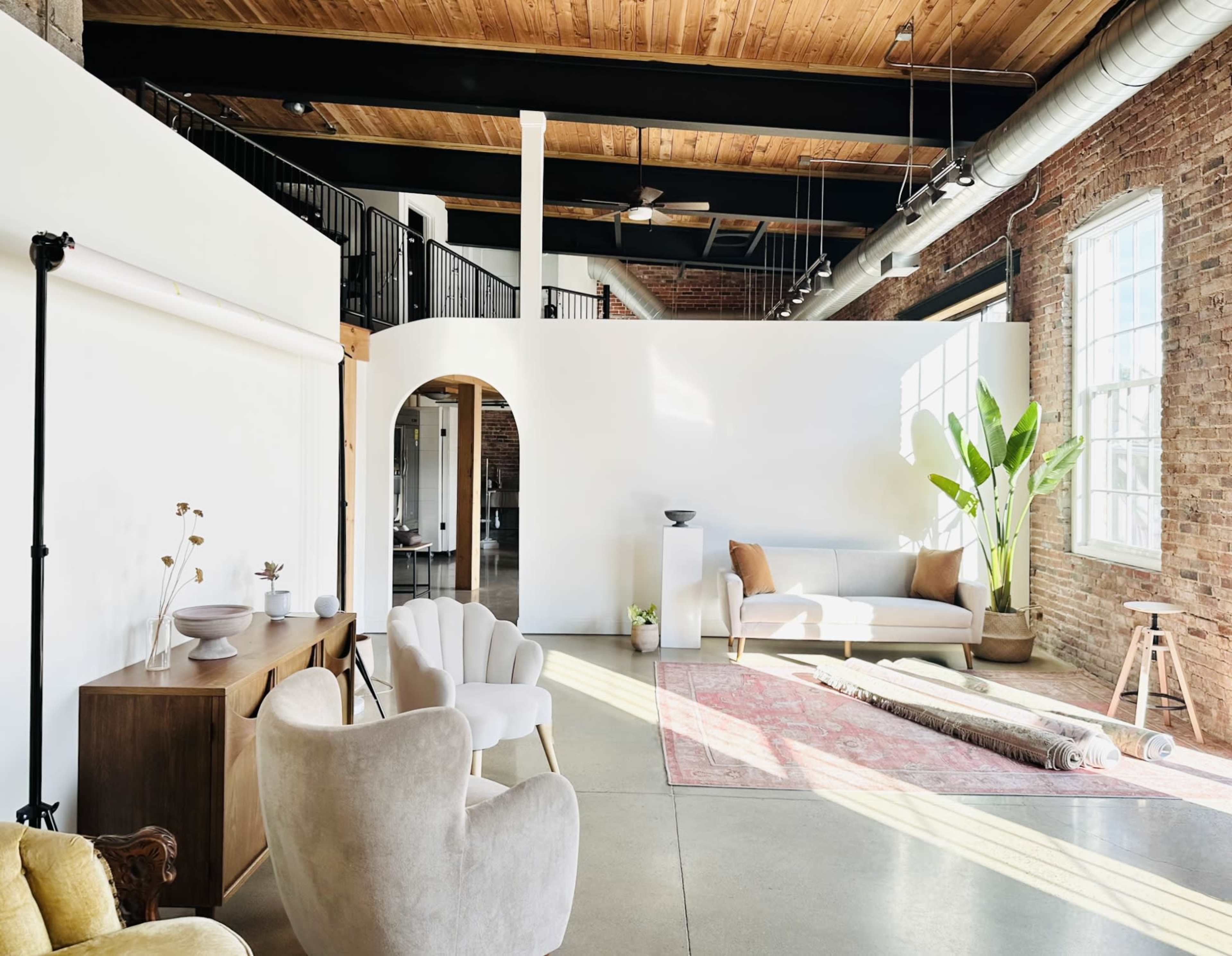 The image shows a spacious, modern interior with exposed brick walls, wooden beams, and a mix of furniture including a sofa, armchairs, and decorative plants.