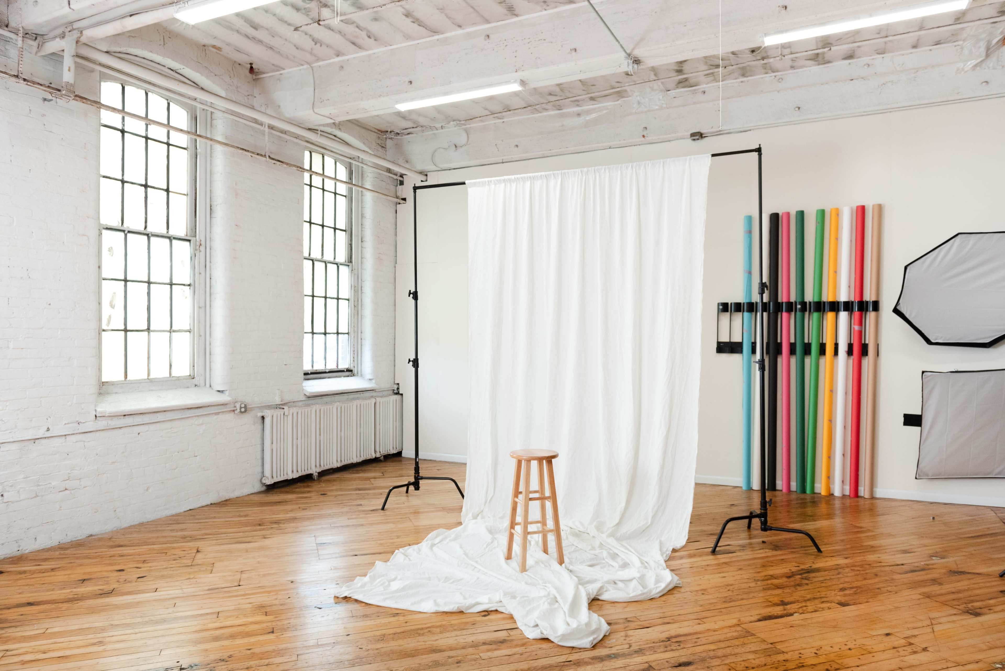 A minimalist photography studio features a wooden stool in front of a white backdrop, with colorful vertical strips on the side walls and large windows allowing natural light.