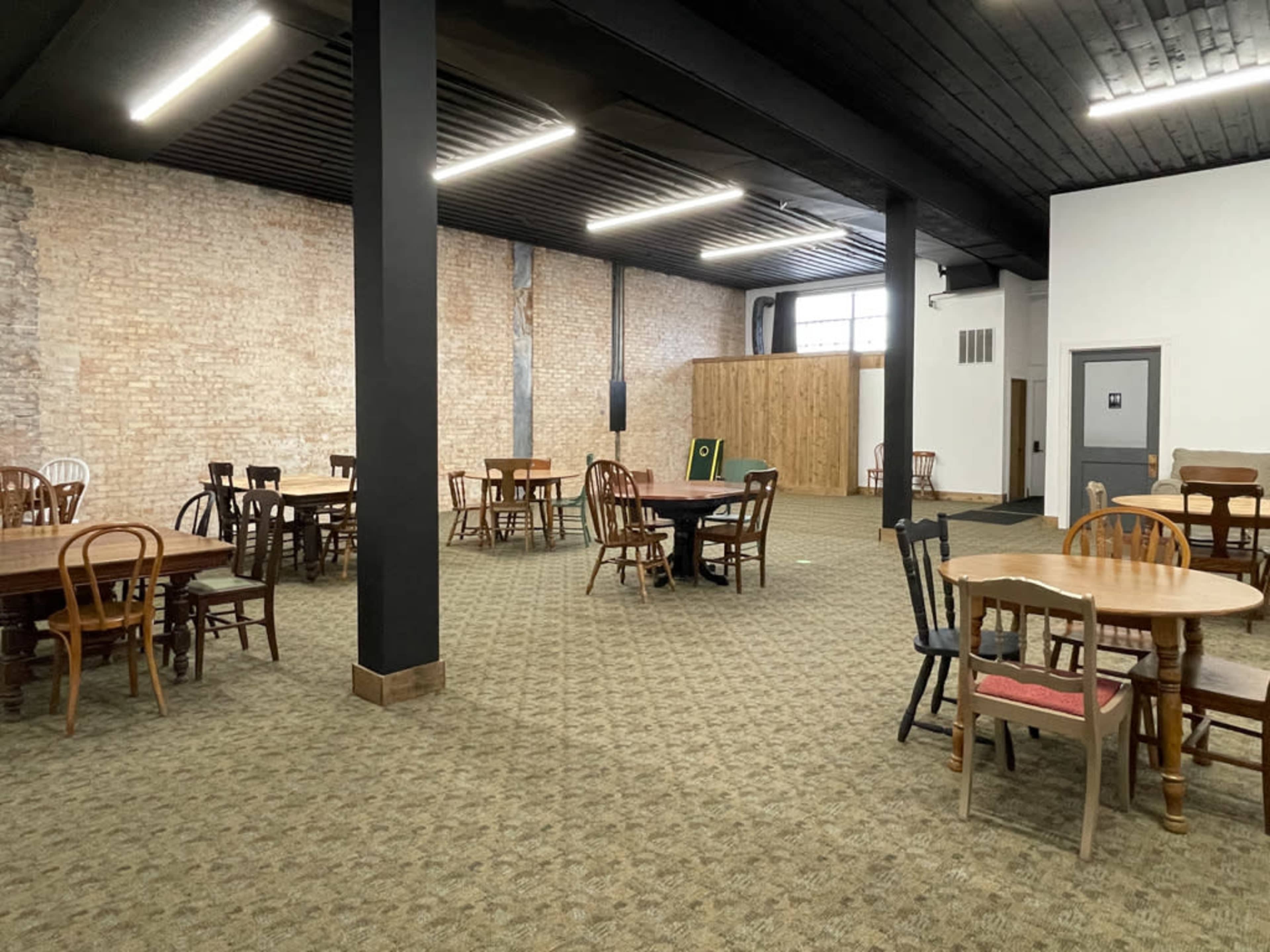 The image shows an empty room with several wooden tables and chairs arranged on a patterned carpet, featuring exposed brick walls and a partitioned area in the background.
