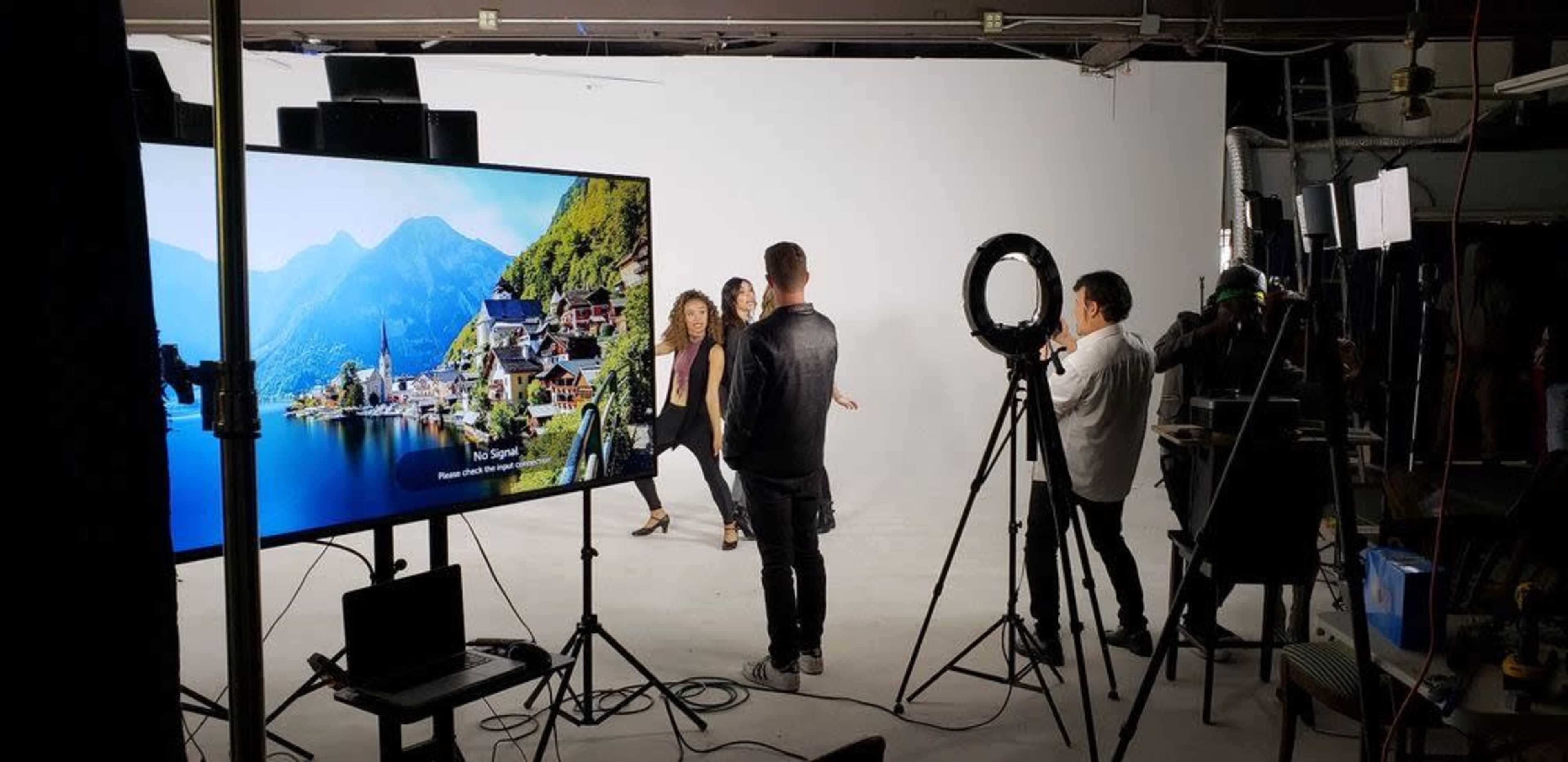 A film crew sets up in a studio with a white backdrop, capturing a scene featuring three individuals in the foreground, while various cameras and lighting equipment are positioned around them.